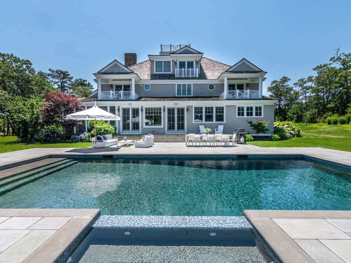 A grand coastal home with balconies and a spacious patio overlooks a sparkling rectangular pool surrounded by lush lawn and trees under a clear blue sky.