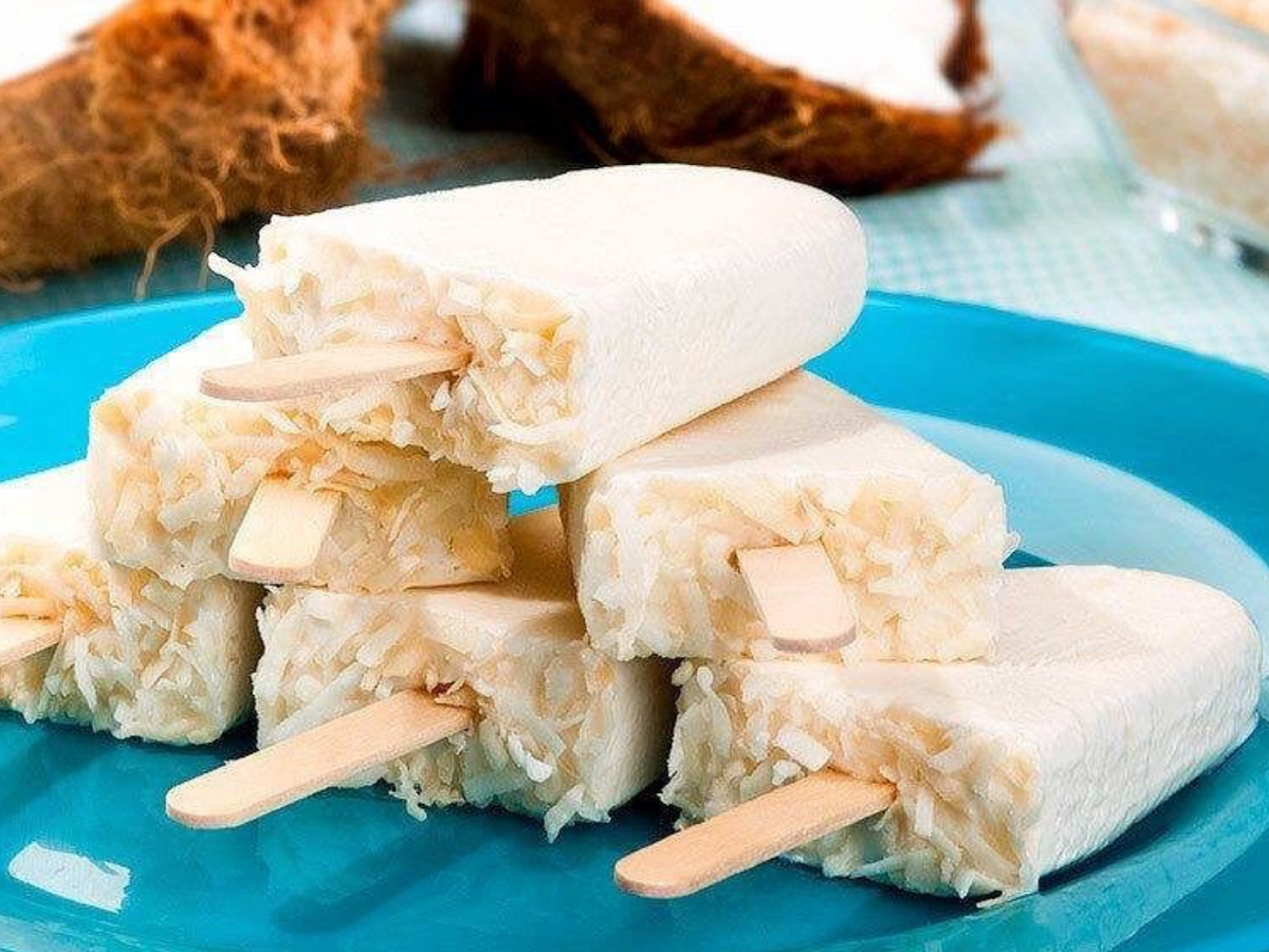 A stack of creamy coconut popsicles with shredded coconut visible inside, arranged on a bright blue plate with halved coconuts in the background.