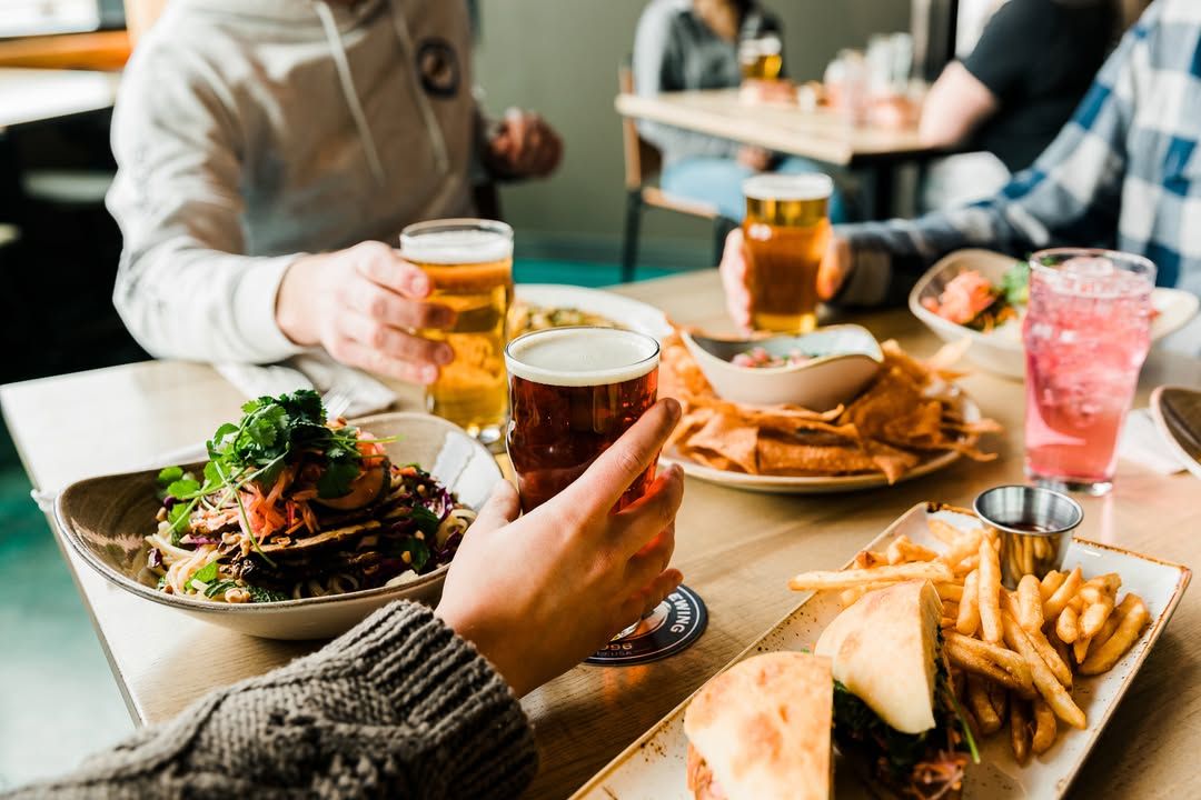 Friends raise craft beer glasses while enjoying burgers and fries at Pelican Brewing near Cannon Beach and Arch Cape. The relaxed pub setting makes it a great stop after a day on the Oregon coast. It’s a well-known brewery for oceanfront dining in Cannon Beach.