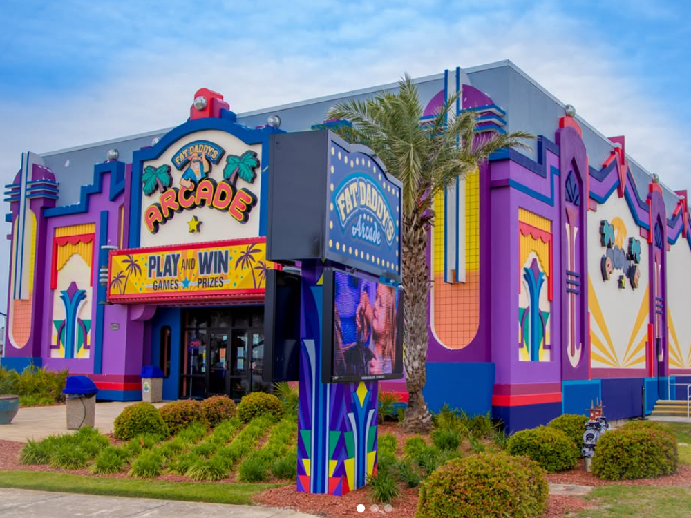 The vibrant exterior of Fat Daddy's Arcade features bold colors, palm trees, and neon signage inviting guests to "Play and Win" games and prizes.