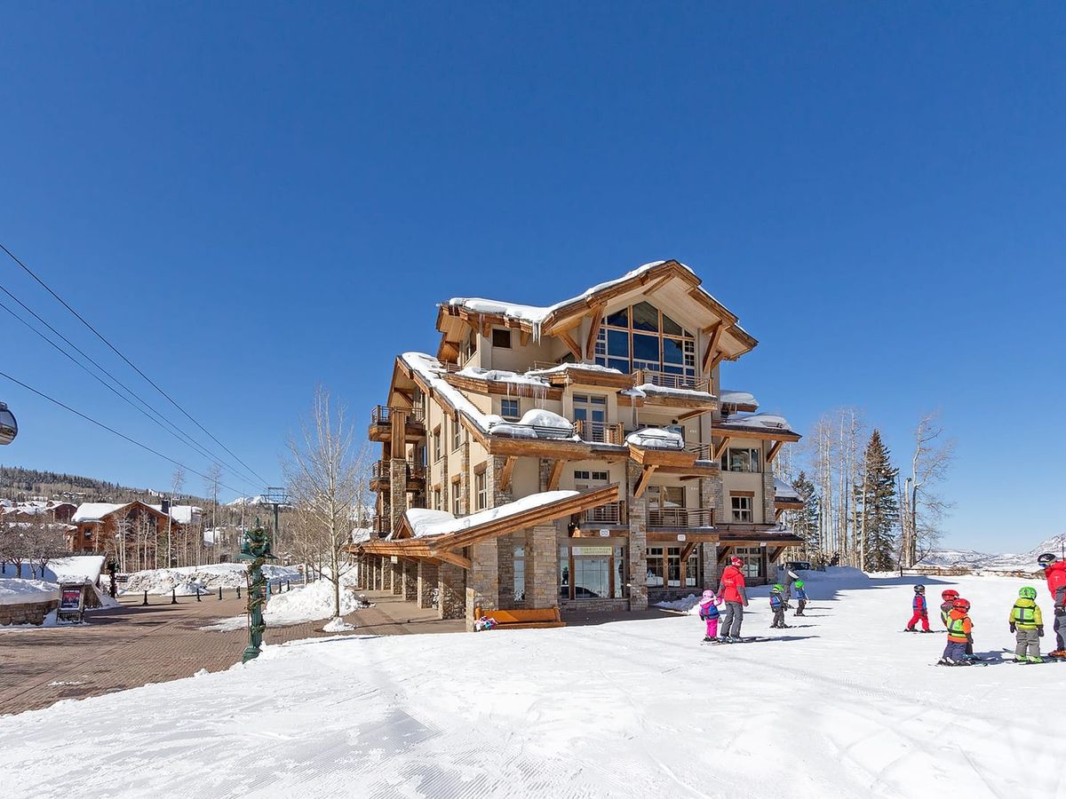 Ski-in ski-out lodge with timber and stone design, snow-covered roof, and children skiing nearby under a clear blue sky.