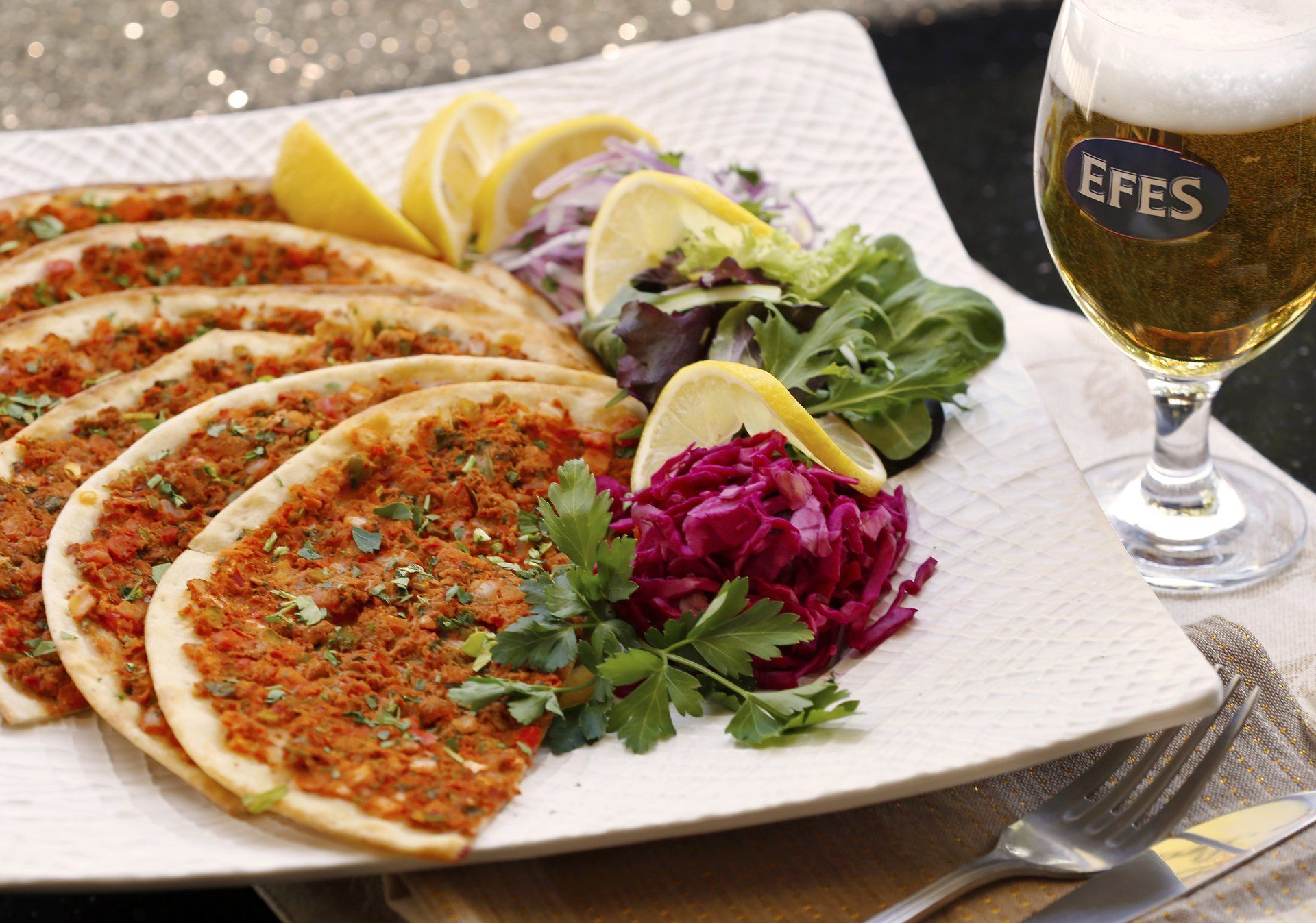 A plate of Turkish lahmacun sits beside fresh herbs, lemon wedges, and colorful cabbage, served with a cold Efes beer. The thin flatbreads are topped with spiced meat and herbs, giving the dish bold color and aroma. It feels like a true taste of Turkey