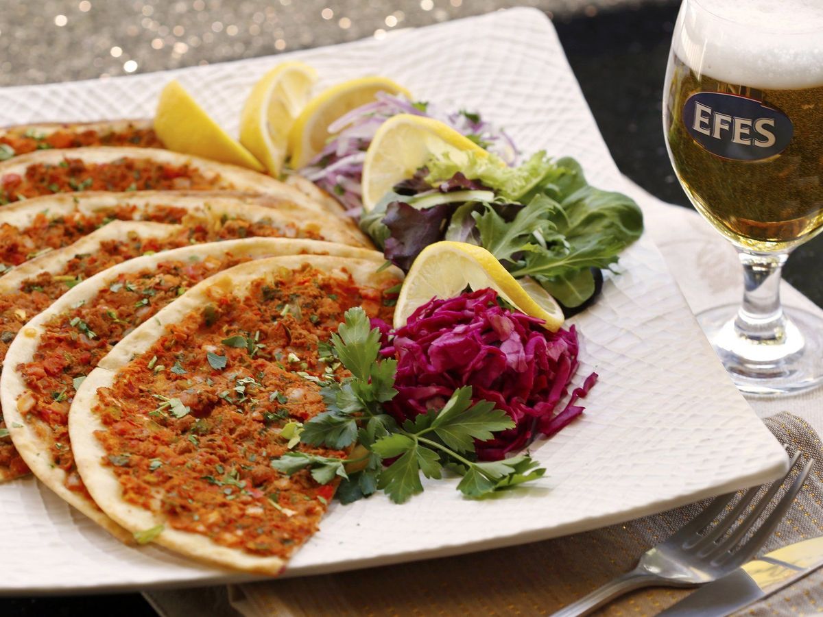 A plate of Turkish lahmacun sits beside fresh herbs, lemon wedges, and colorful cabbage, served with a cold Efes beer. The thin flatbreads are topped with spiced meat and herbs, giving the dish bold color and aroma. It feels like a true taste of Turkey