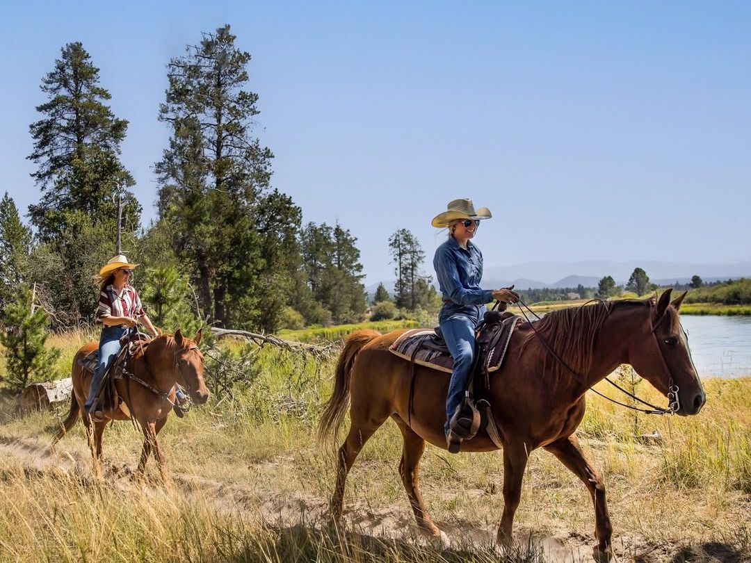 Two people enjoy horseback riding along a river trail surrounded by open fields and pine trees. Horseback adventures in Sunriver let you experience the region’s rugged beauty and western spirit up close.