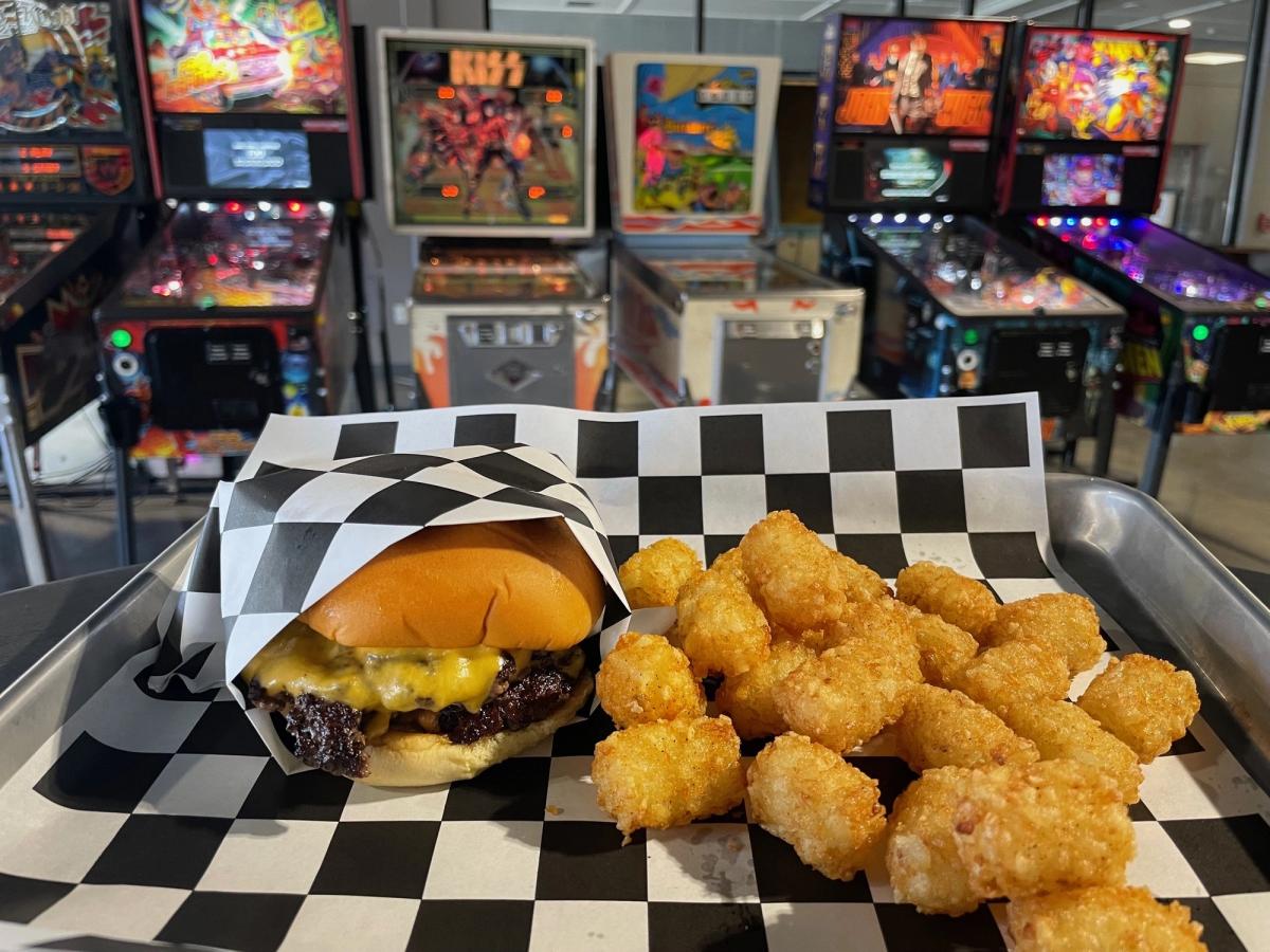 Cheeseburger and crispy tater tots served on a checkered tray in front of a row of vintage pinball machines. Perfect mix of comfort food and retro arcade fun.