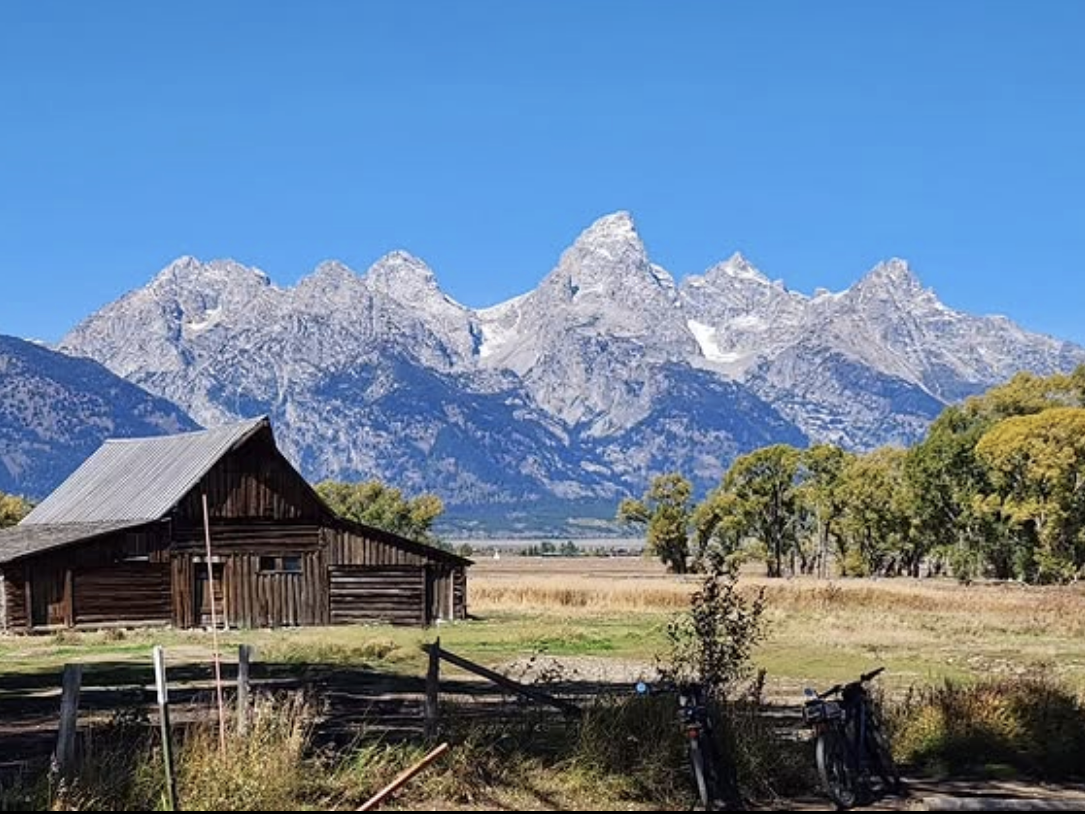 Historic wooden barn set in a golden field with a backdrop of snow-dusted mountain peaks and clear blue skies.
