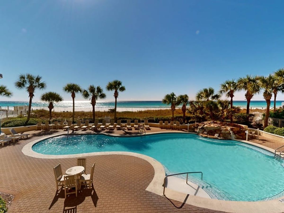 A large beachfront pool overlooks the emerald waters of Destin, Florida, with palm trees lining the oceanfront deck. Lounge chairs sit beside the pool, offering clear views of the Gulf Coast. Destin is famous for its sugar-white sand and family-friendly beach resorts.
