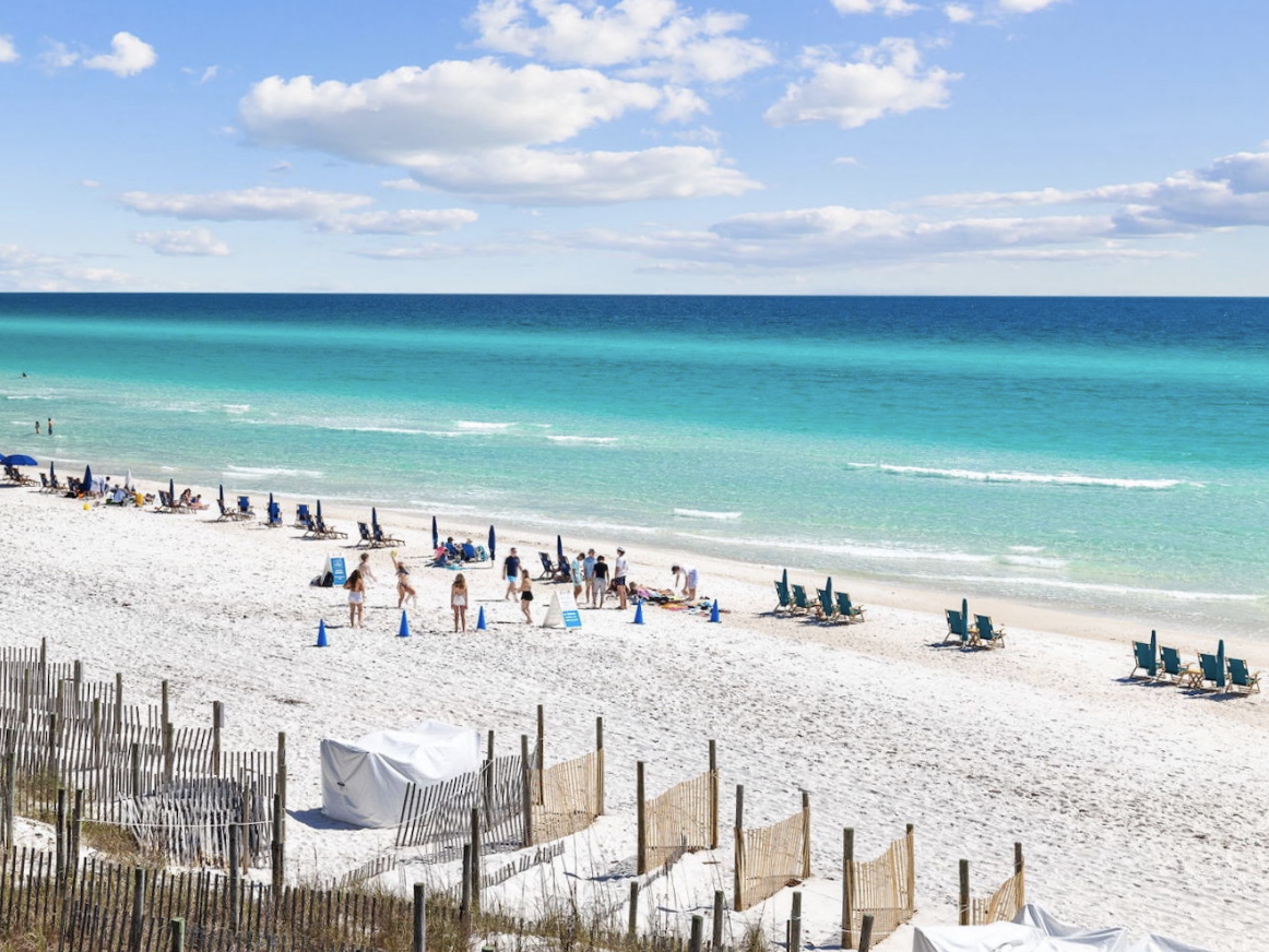 White sandy beach with rows of teal lounge chairs and umbrellas, groups of people enjoying the sun and sea, and turquoise waves sparkling under a bright sky with scattered clouds.