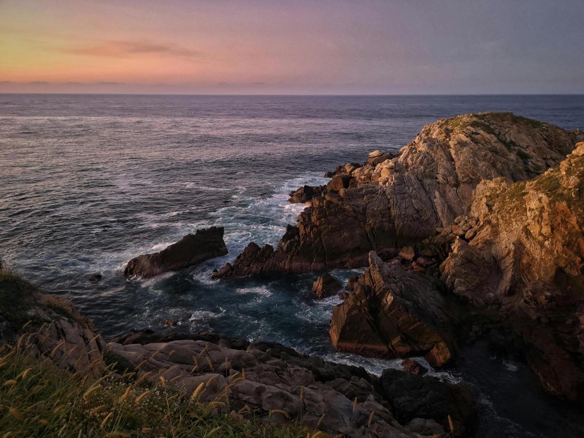Waves crash against rugged cliffs in Huatulco, Mexico, as the sky glows with soft pink and orange hues at sunset. The dramatic shoreline shows the beauty of Oaxaca’s Pacific coast.