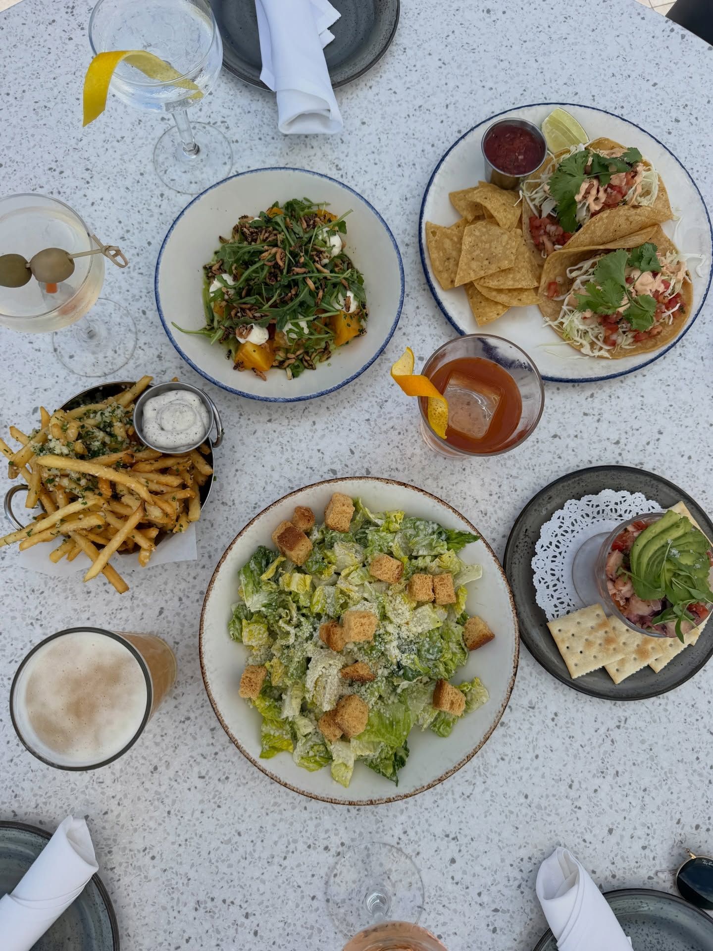 An overhead spread of coastal dishes covers a terrazzo table, including shrimp tacos with chips and salsa, a Caesar salad, a golden beet salad, garlic parmesan fries, and ceviche topped with avocado. Craft cocktails and a beer round out the bright, breezy seaside meal.
