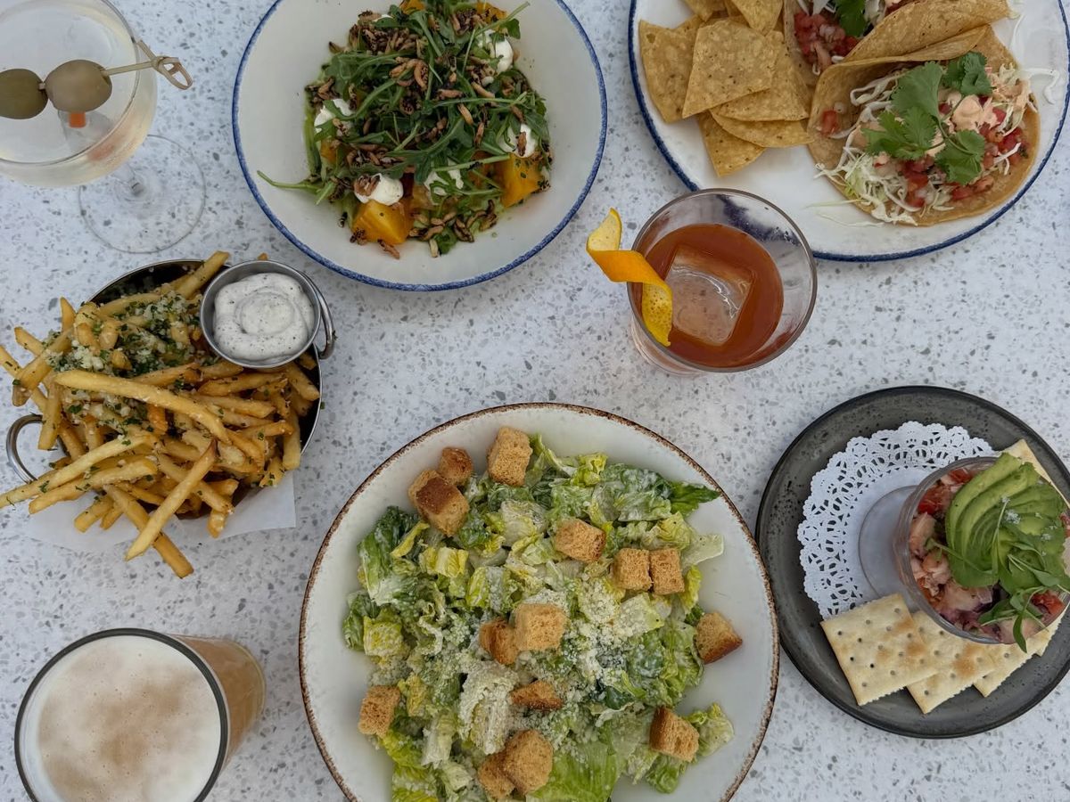 An overhead spread of coastal dishes covers a terrazzo table, including shrimp tacos with chips and salsa, a Caesar salad, a golden beet salad, garlic parmesan fries, and ceviche topped with avocado. Craft cocktails and a beer round out the bright, breezy seaside meal.
