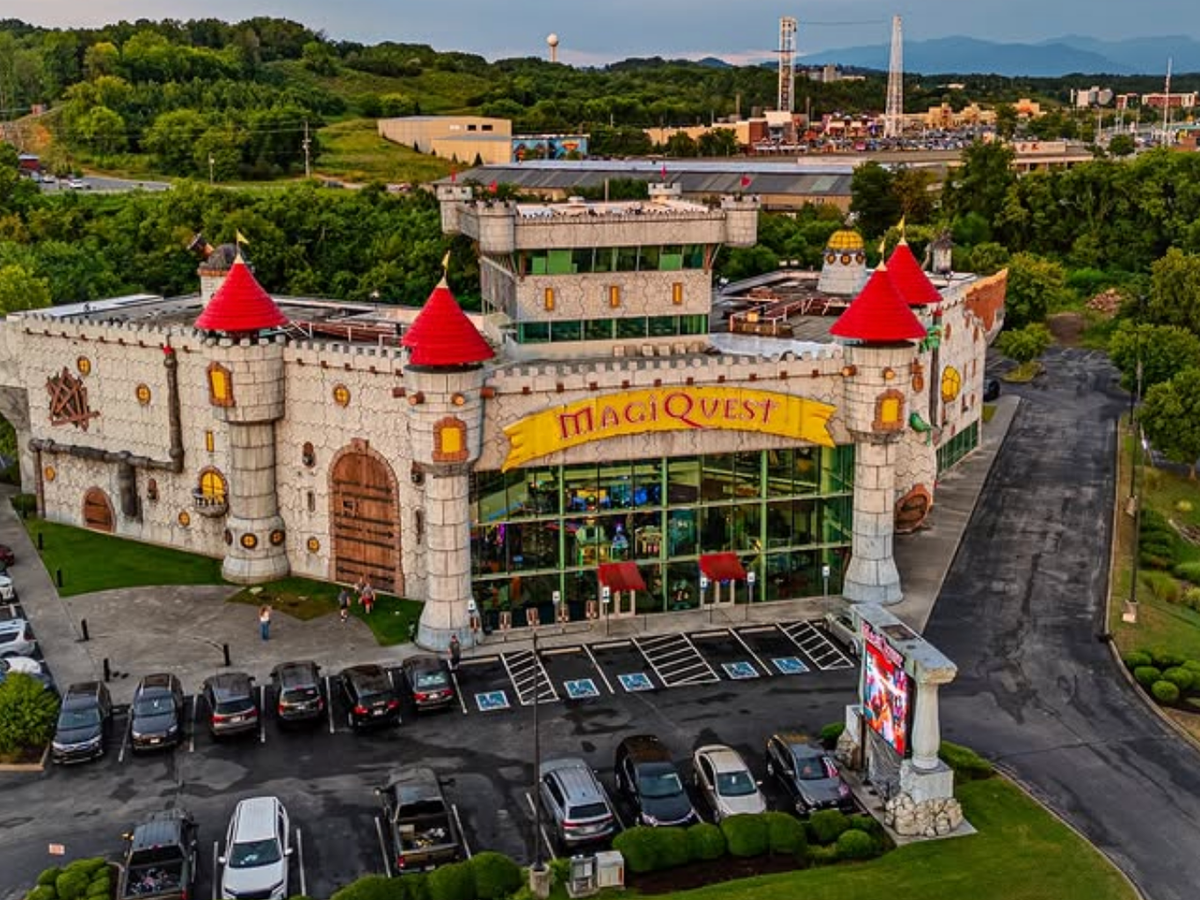 A whimsical castle-themed building with red turrets and stone-like walls houses MagiQuest, with colorful banners, large wooden doors, and a parking lot filled with cars in front.