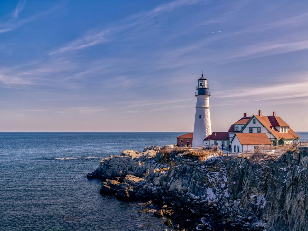 A classic white lighthouse with a black top stands on a rocky cliffside overlooking the ocean, accompanied by red-roofed buildings under a vibrant blue sky.