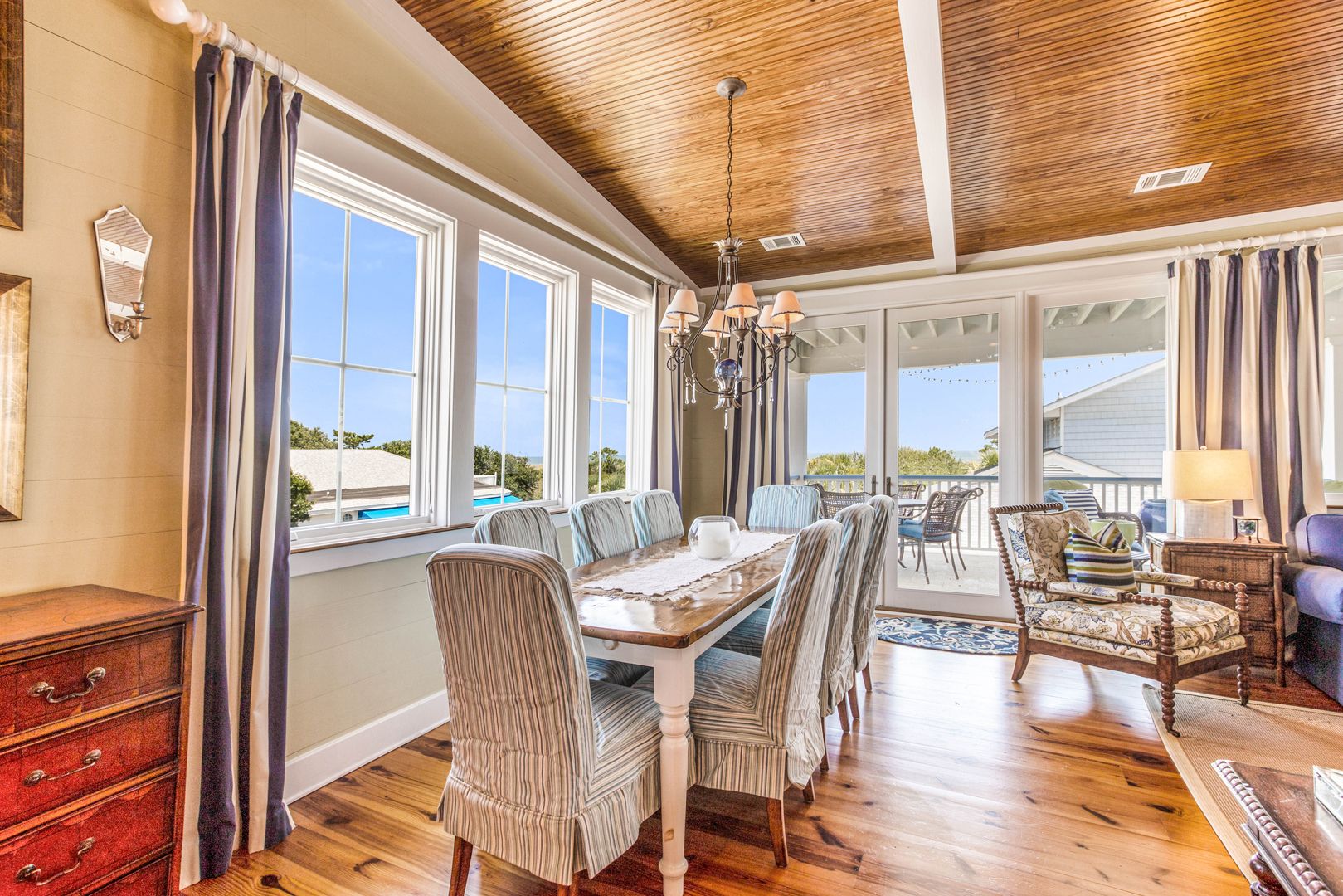 Bright coastal dining room with a wooden table, striped slipcovered chairs, and a chandelier, featuring large windows and glass doors that open to a covered balcony with outdoor seating.