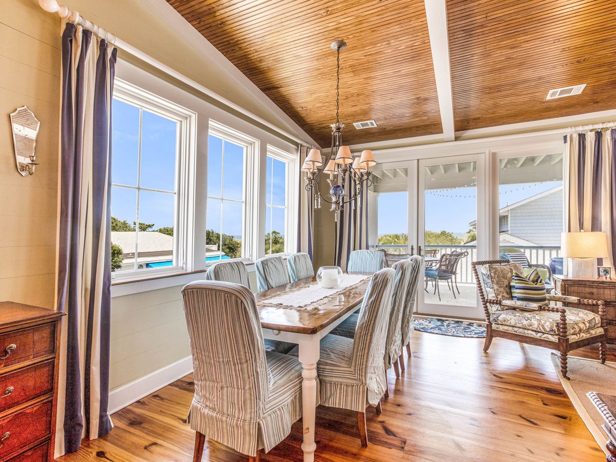 Bright coastal dining room with a wooden table, striped slipcovered chairs, and a chandelier, featuring large windows and glass doors that open to a covered balcony with outdoor seating.