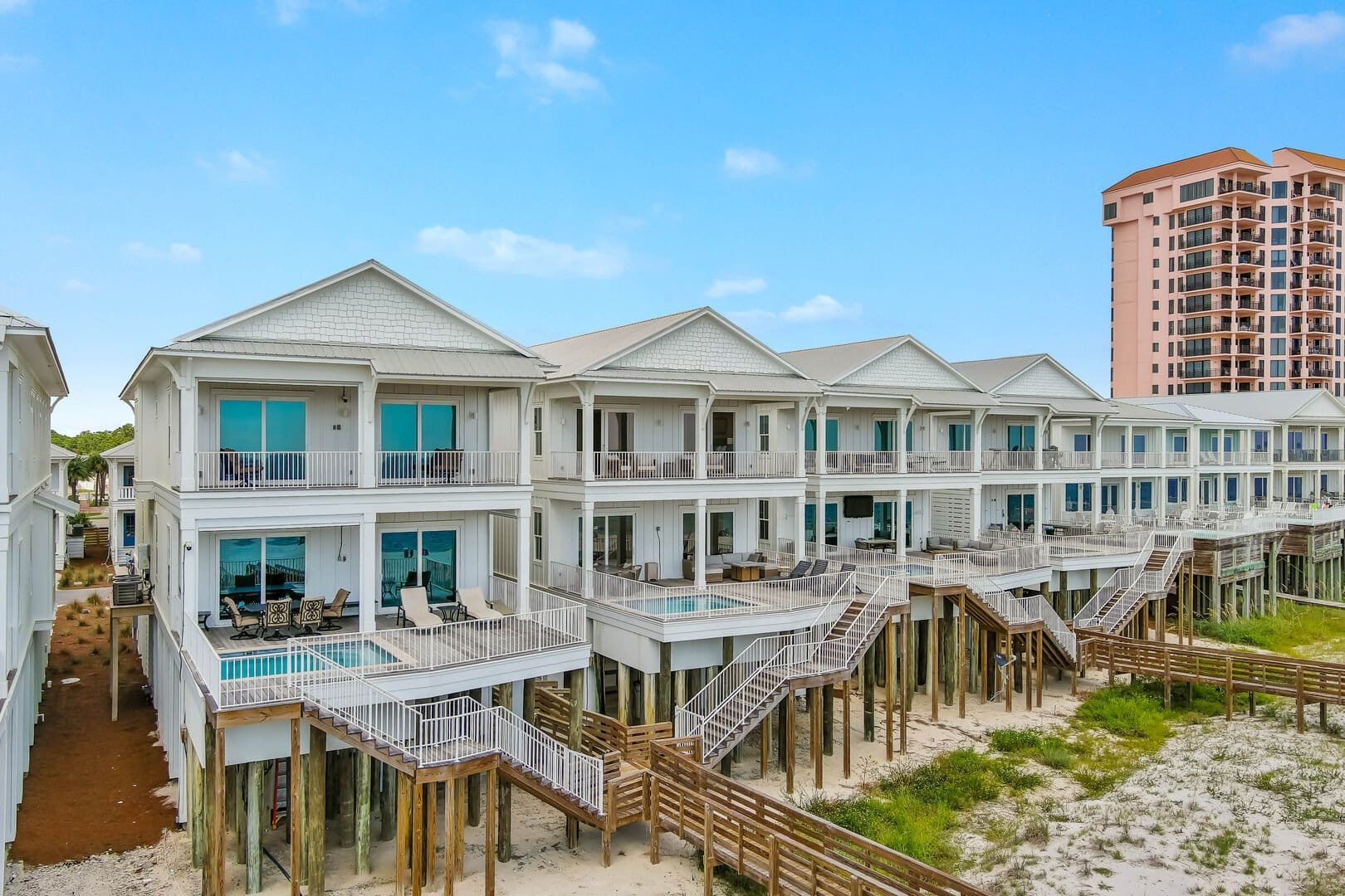 Row of elevated beachfront vacation homes with private balconies, pools, and direct stair access to the sand. The homes feature a coastal white exterior with large windows overlooking the ocean.