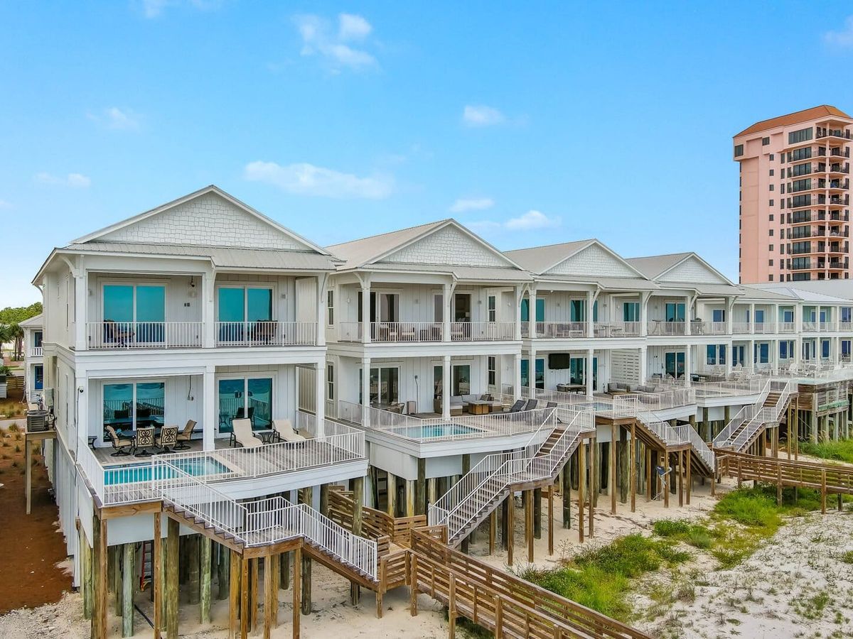 Row of elevated beachfront vacation homes with private balconies, pools, and direct stair access to the sand. The homes feature a coastal white exterior with large windows overlooking the ocean.