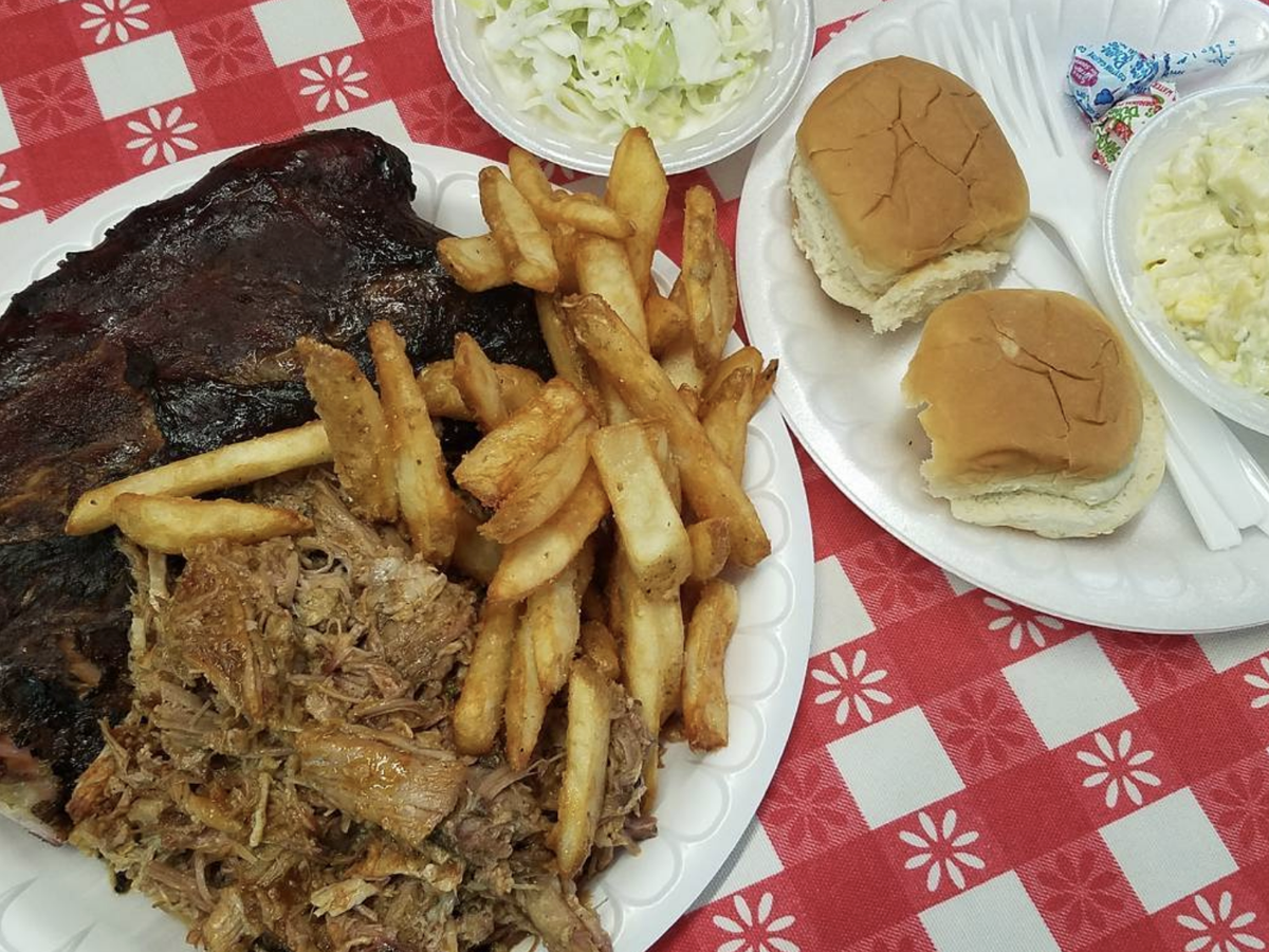 A classic BBQ meal on a red checkered tablecloth, featuring a plate of ribs, pulled pork, and seasoned fries, accompanied by bowls of coleslaw and potato salad, and two soft dinner rolls.