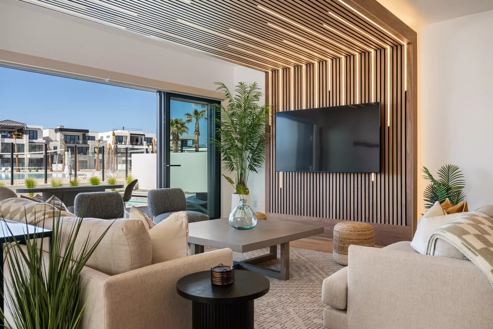 Modern living room with neutral-toned seating, a wood-slat accent wall with mounted TV, and large glass doors opening to a poolside view.
