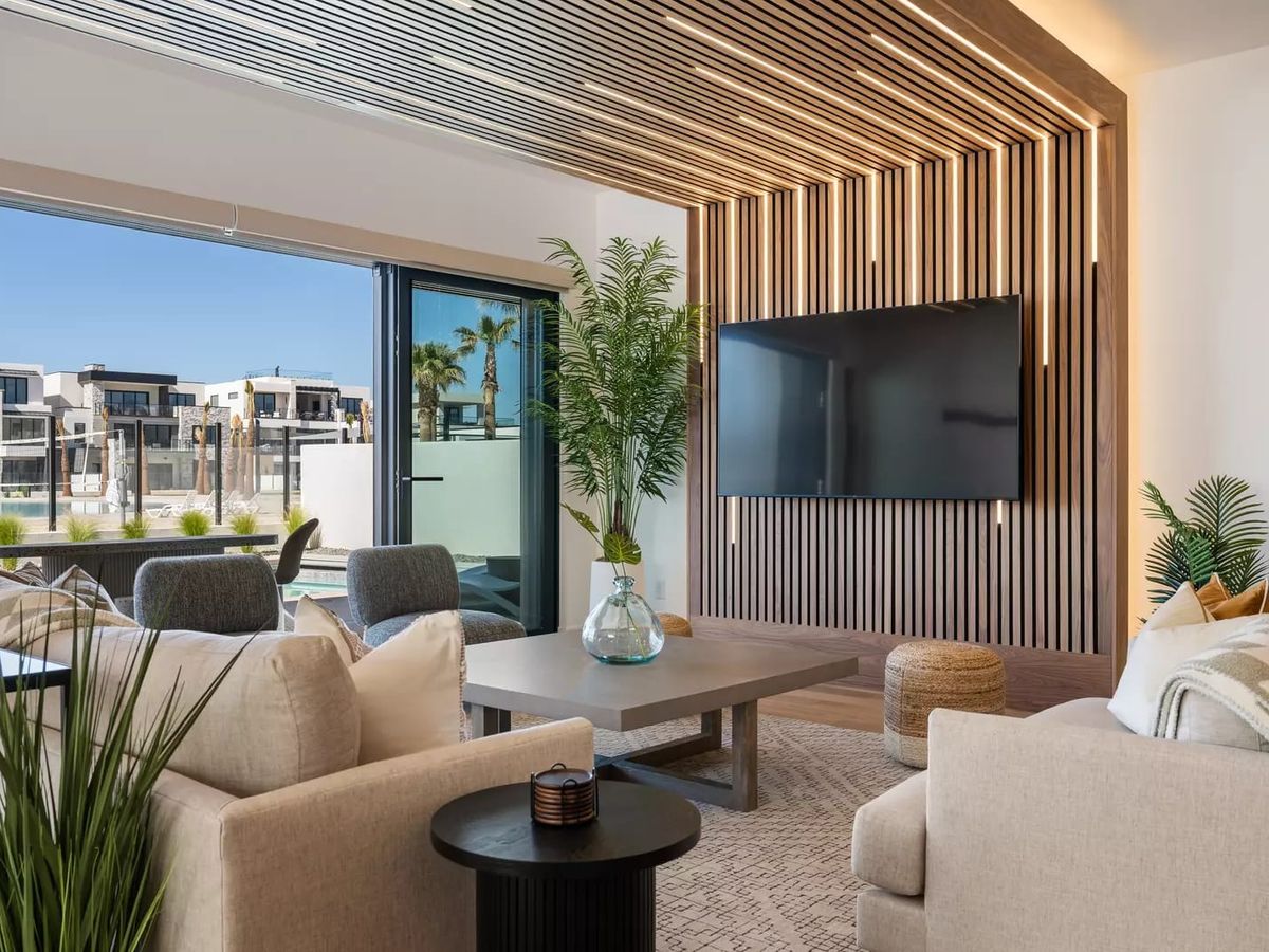 Modern living room with neutral-toned seating, a wood-slat accent wall with mounted TV, and large glass doors opening to a poolside view.