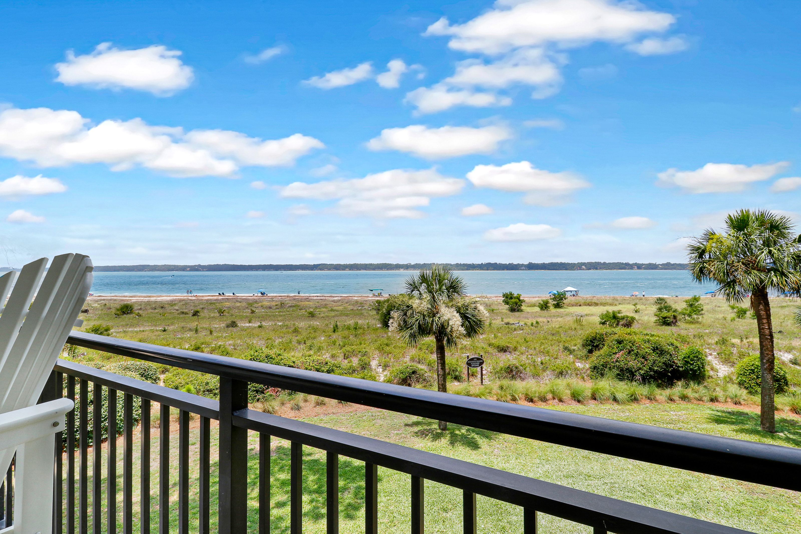 A scenic oceanfront view from a balcony with white Adirondack chairs overlooks grassy dunes, palm trees, and a sandy beach leading to calm blue waters under a bright sky with scattered clouds.