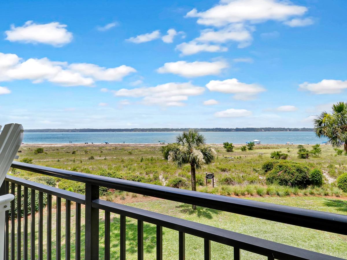 A scenic oceanfront view from a balcony with white Adirondack chairs overlooks grassy dunes, palm trees, and a sandy beach leading to calm blue waters under a bright sky with scattered clouds.