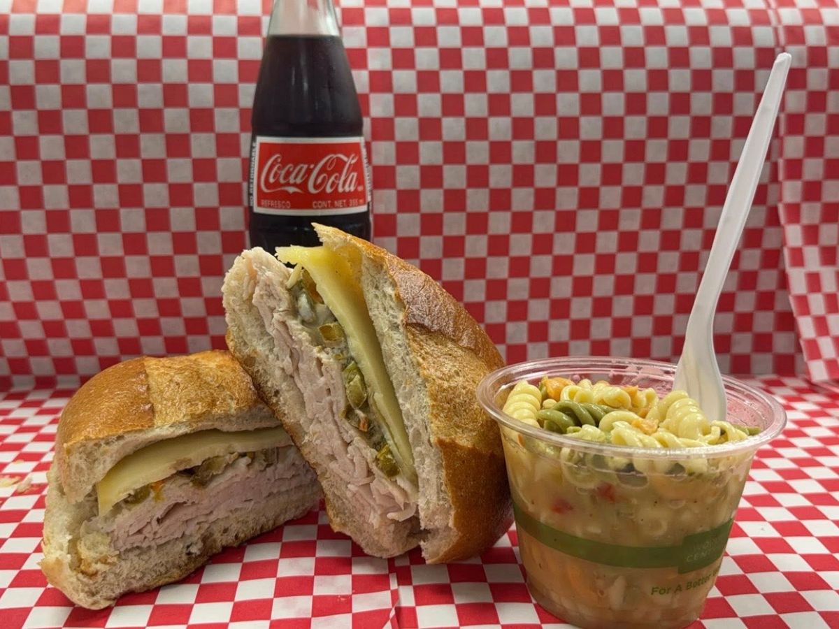 Stacked turkey sandwich with melted cheese and chopped pickles, served with a side of tri-color pasta salad and a glass bottle of Coke. Classic deli-style lunch with bold flavors and a retro vibe.