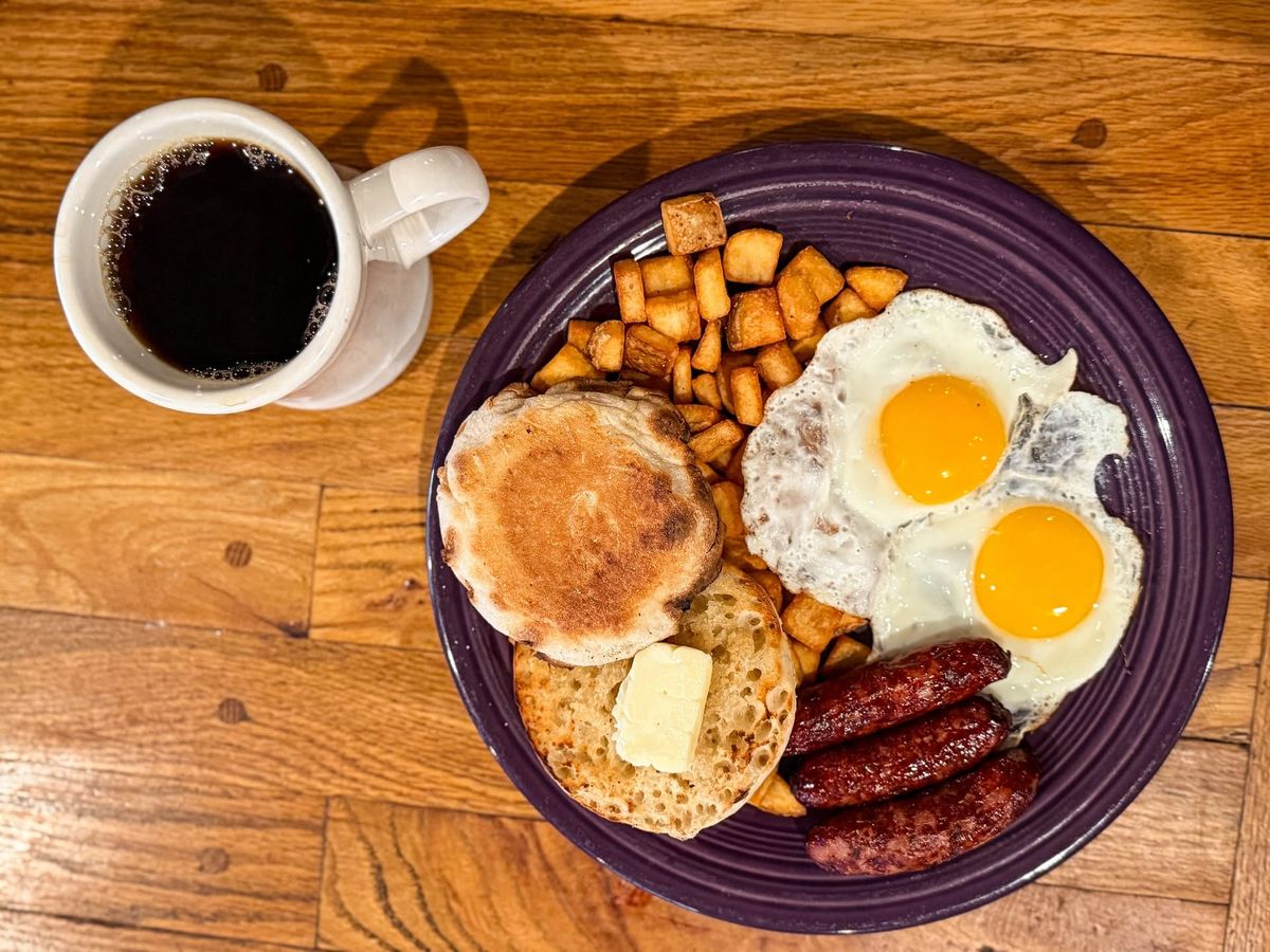 A hearty breakfast plate holds two sunny-side-up eggs, crispy home fries, browned sausage links, and a buttered English muffin on a deep purple plate. A steaming mug of black coffee sits beside it on the warm wooden table.