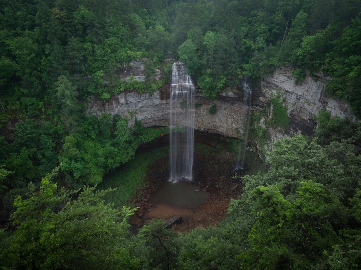 A tall, narrow waterfall cascades over a rocky cliff into a lush, green basin surrounded by dense forest and steep canyon walls.
