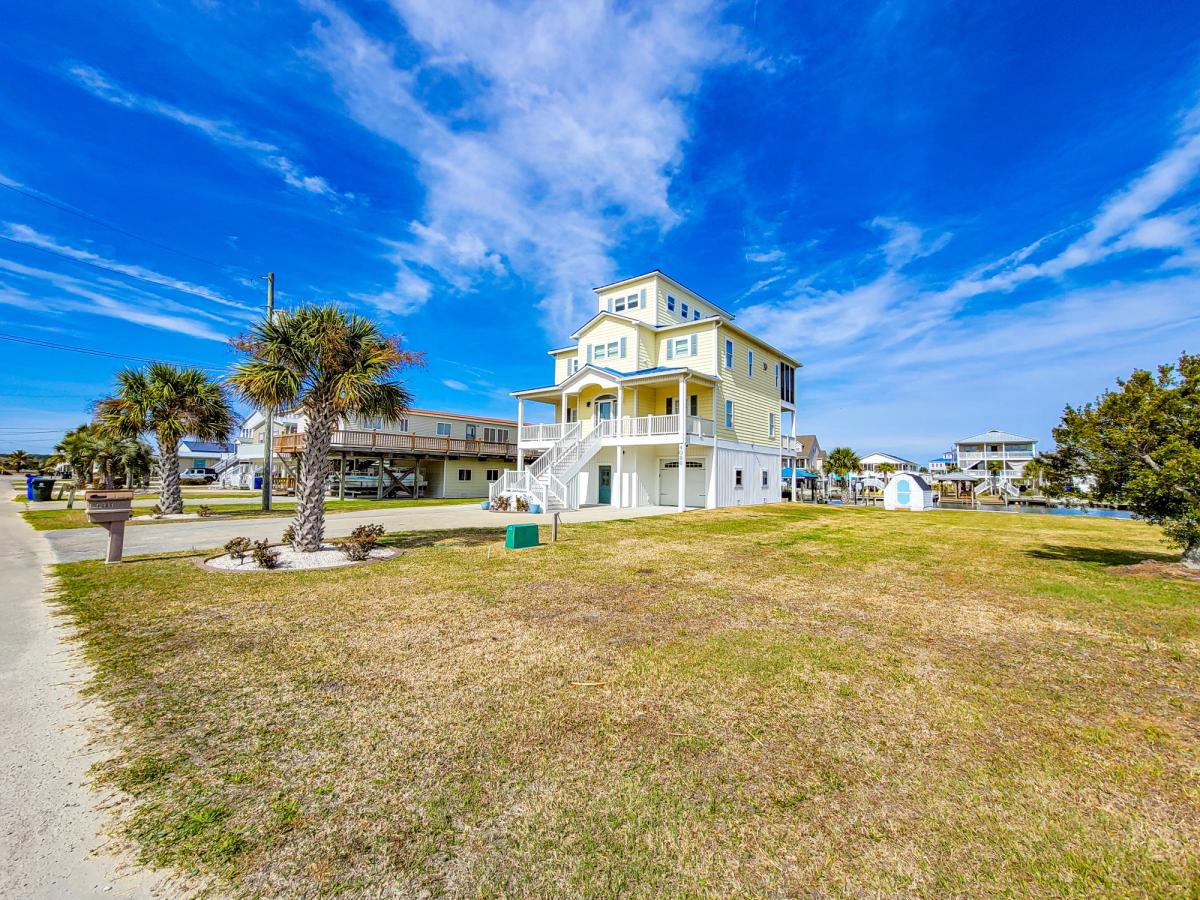 A bright yellow three-story coastal home with white trim and a spacious lawn, surrounded by palm trees and other beach houses under a vivid blue sky with wispy clouds.
