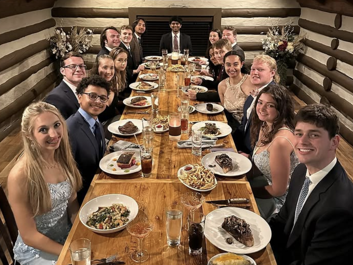 A large group of well-dressed young adults seated around a long wooden table in a cozy log cabin-style dining room, enjoying a formal dinner with plated entrees, drinks, and elegant decor.