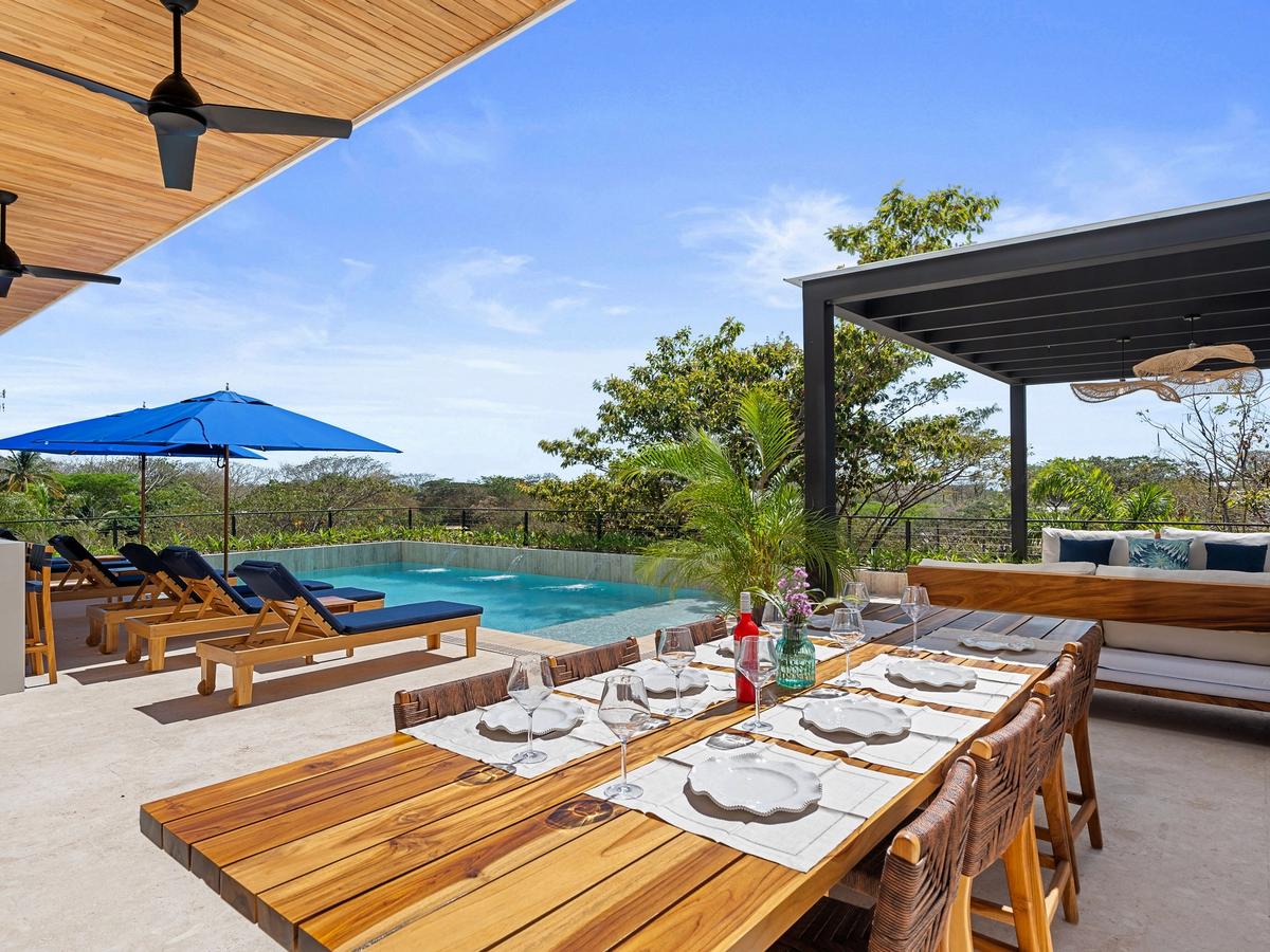 A modern outdoor patio with a wooden dining table set for six, lounge chairs under blue umbrellas, and a private infinity pool surrounded by lush greenery.