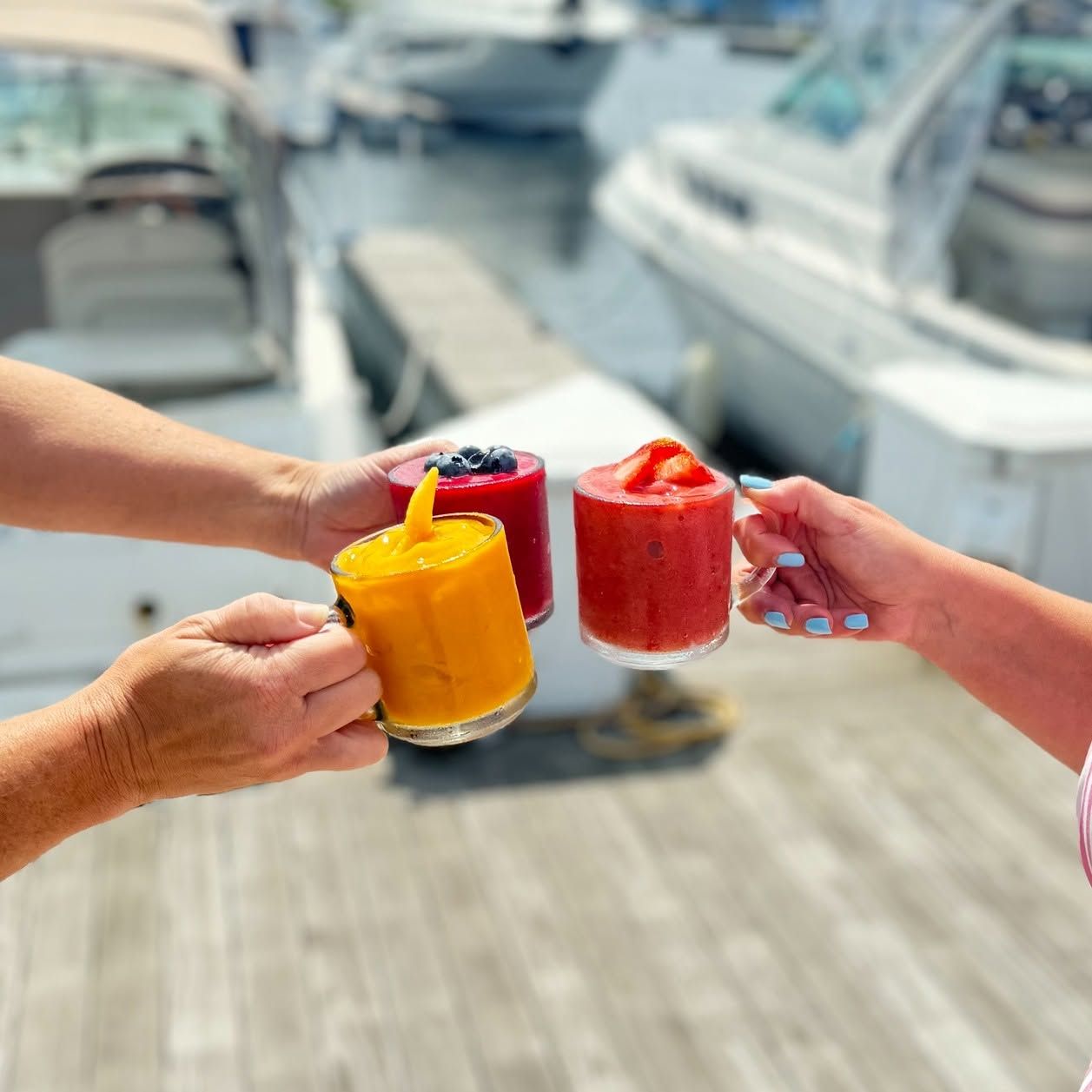 Three hands raise colorful frozen fruit cocktails garnished with berries and strawberries in a toast above a sunlit marina dock. Boats rest in their slips in the background, capturing a bright, festive waterfront moment.