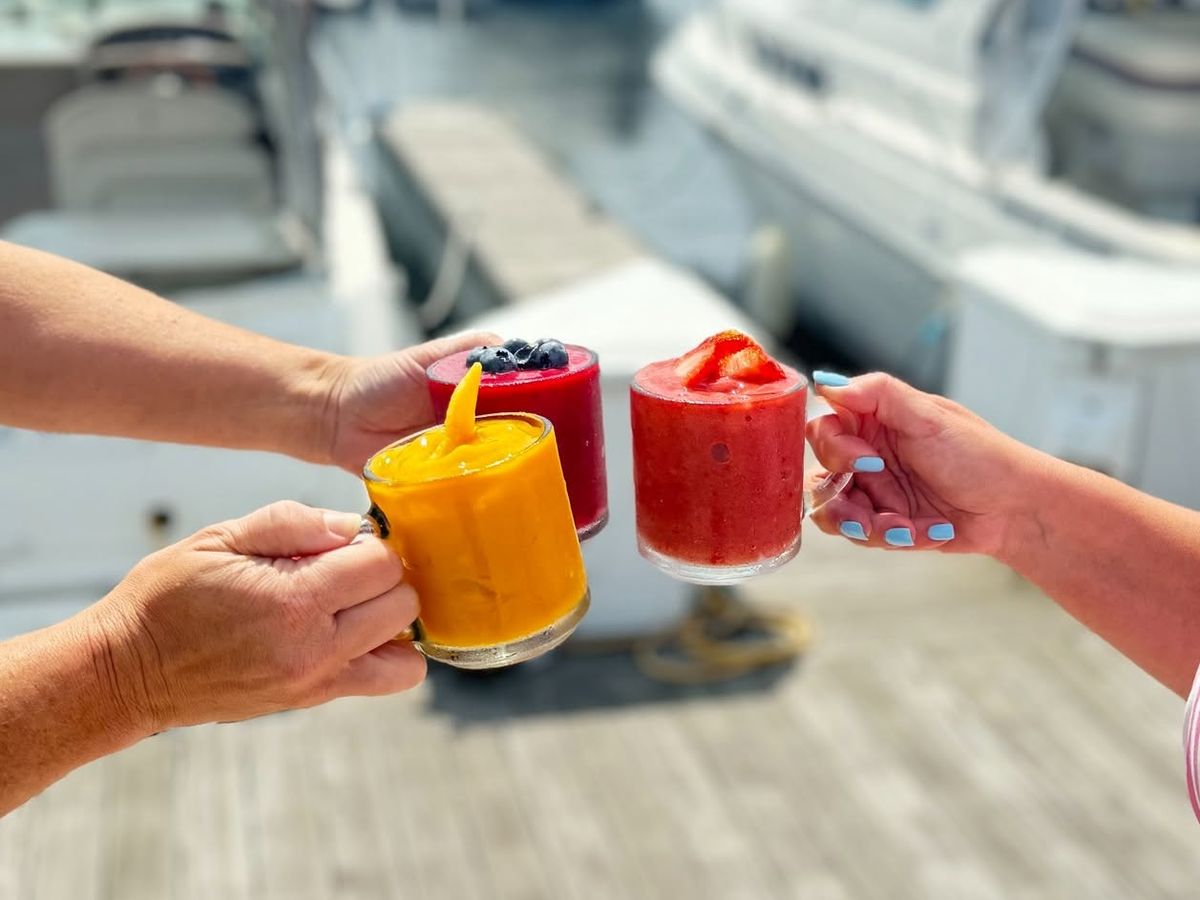 Three hands raise colorful frozen fruit cocktails garnished with berries and strawberries in a toast above a sunlit marina dock. Boats rest in their slips in the background, capturing a bright, festive waterfront moment.