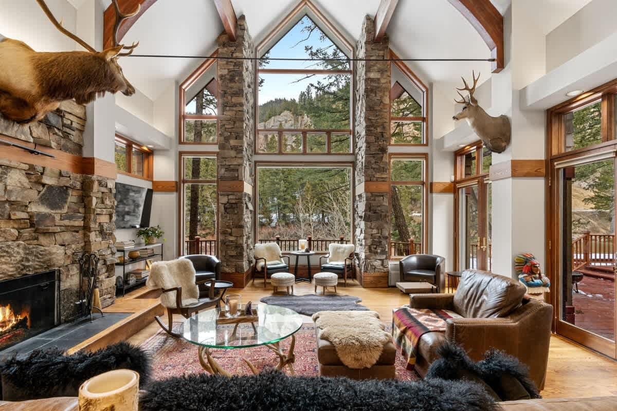 A grand mountain lodge living room with soaring windows, stone columns, and a fireplace features leather seating, fur throws, and forest views framed by the glass.