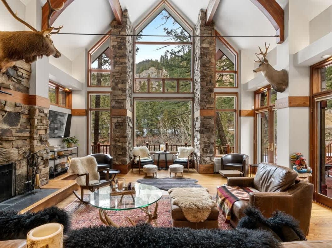A grand mountain lodge living room with soaring windows, stone columns, and a fireplace features leather seating, fur throws, and forest views framed by the glass.