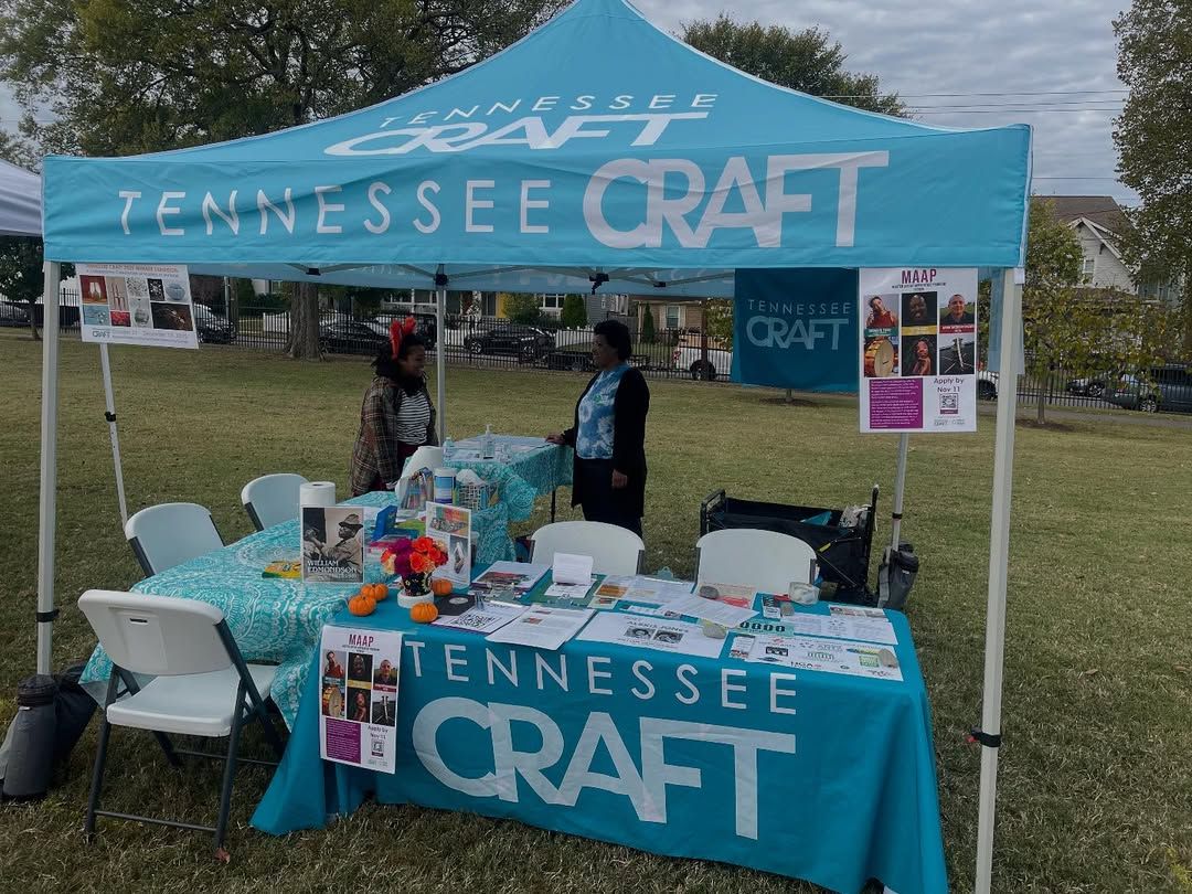 A bright blue Tennessee Craft tent sits on a grassy field with friendly volunteers chatting at the booth. The table is filled with flyers, artwork, and small displays that invite people to learn more. It feels like a relaxed community event where everyone stops by to explore local art.