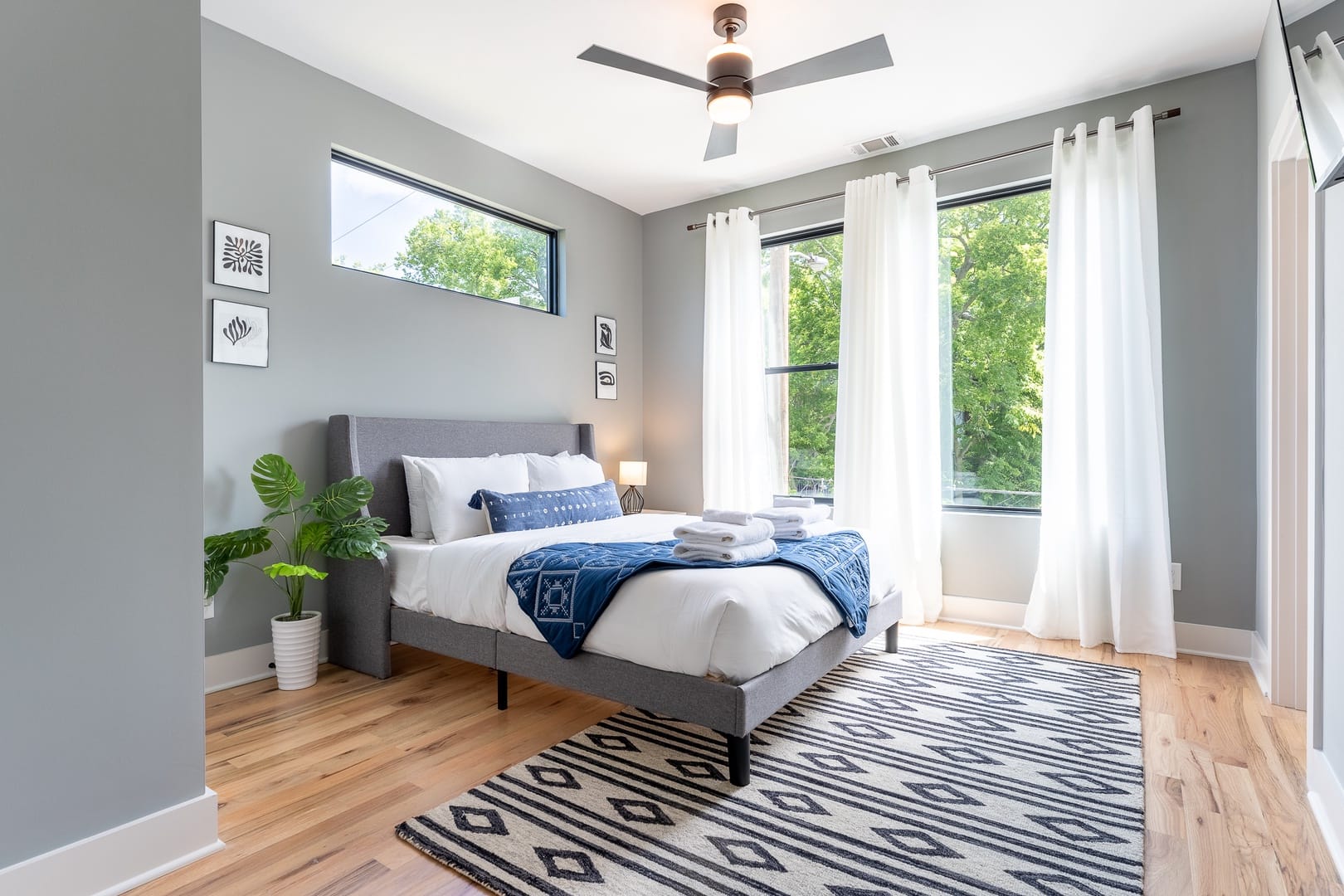 A bright and airy bedroom with a gray upholstered bed, crisp white linens, blue accents, and large windows framed by sheer white curtains overlooking greenery.