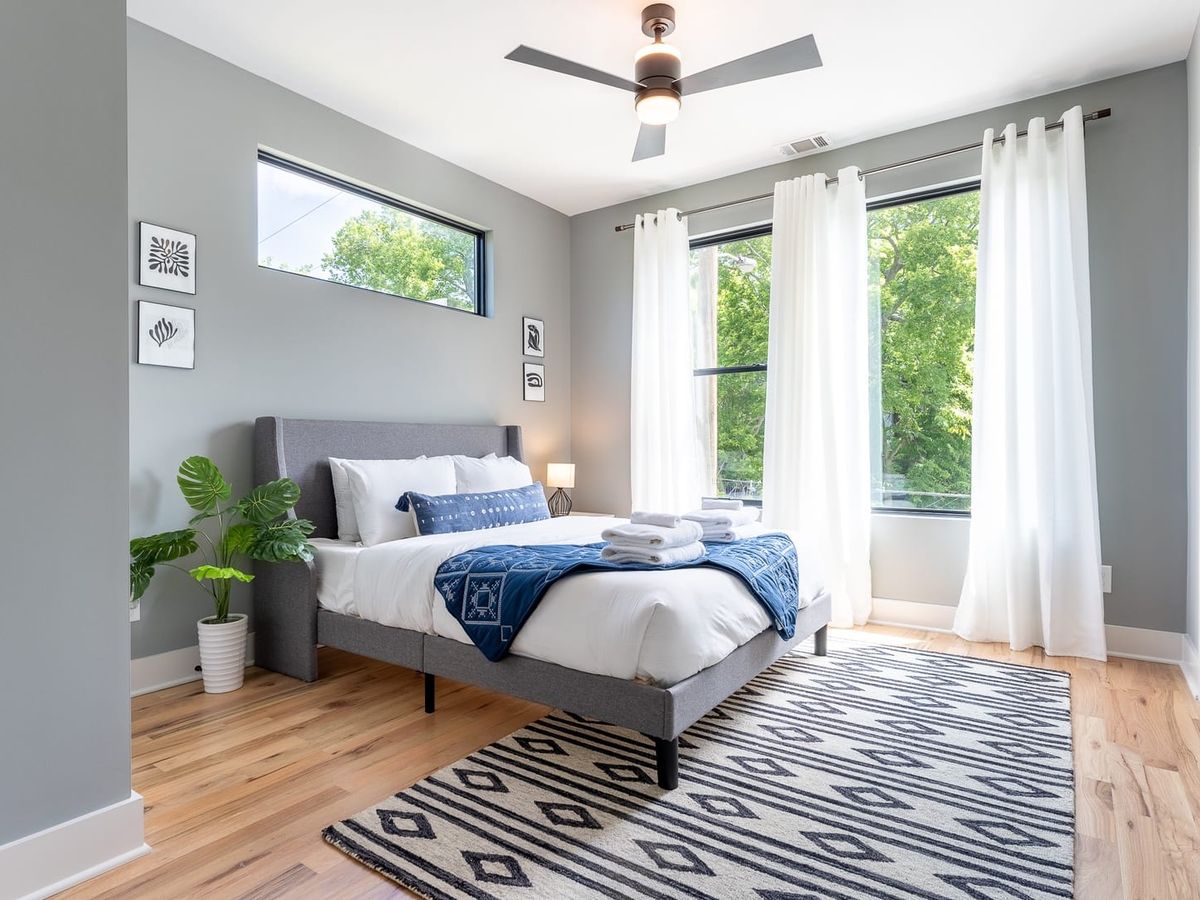 A bright and airy bedroom with a gray upholstered bed, crisp white linens, blue accents, and large windows framed by sheer white curtains overlooking greenery.