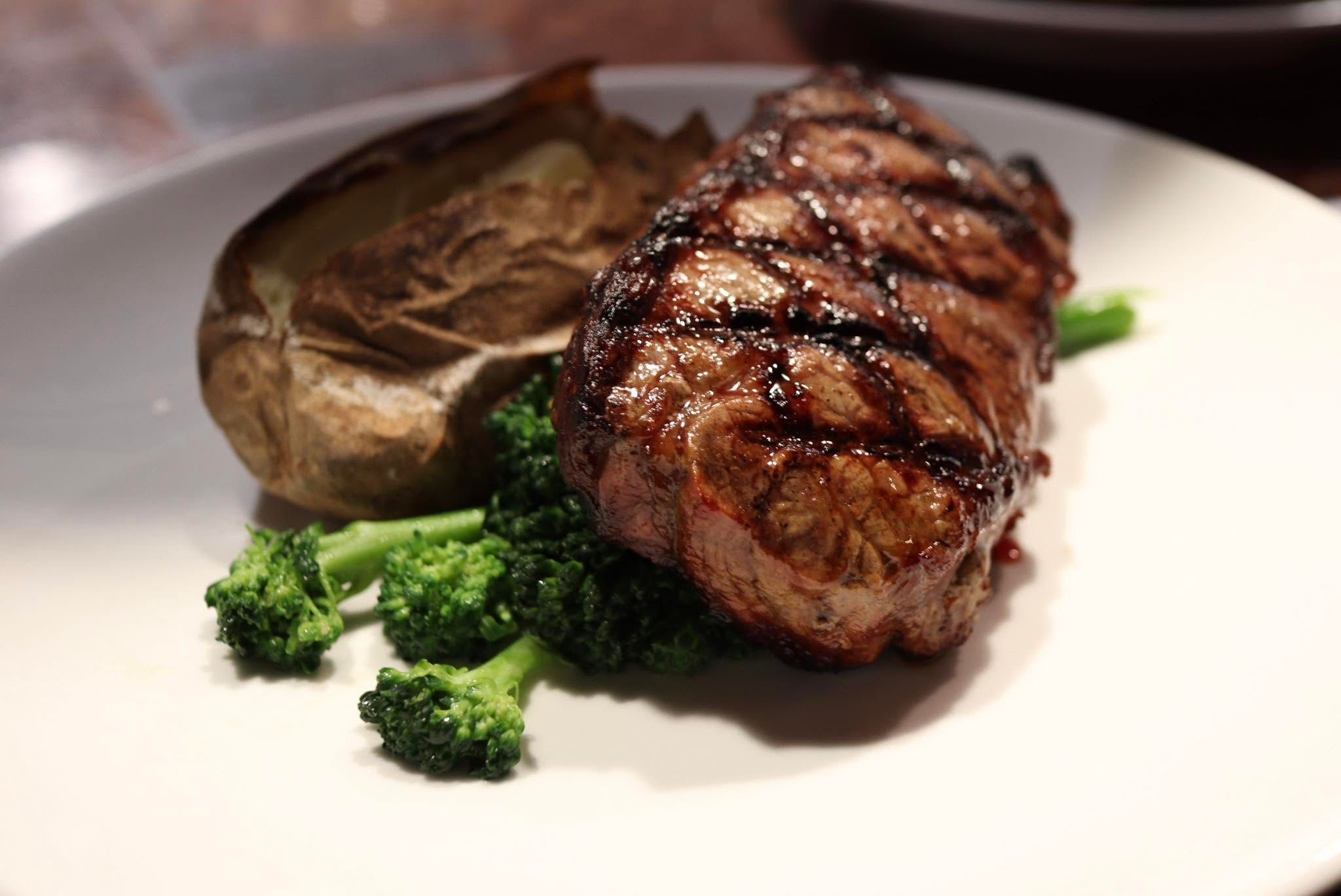 A thick, char-grilled steak with deep crosshatch grill marks rests against a foil-wrapped baked potato and a cluster of steamed broccoli on a white plate. Warm lighting emphasizes the juicy, perfectly seared cut of beef.