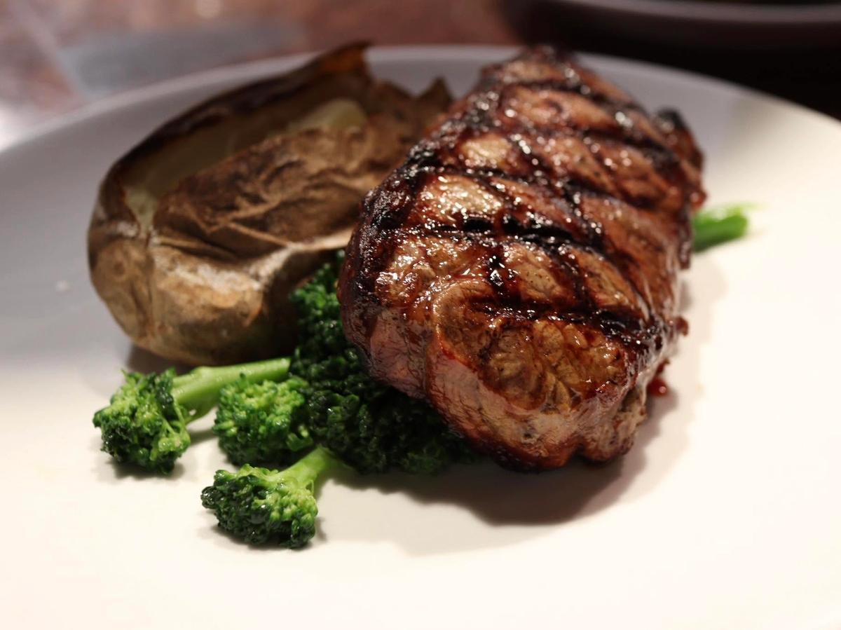 A thick, char-grilled steak with deep crosshatch grill marks rests against a foil-wrapped baked potato and a cluster of steamed broccoli on a white plate. Warm lighting emphasizes the juicy, perfectly seared cut of beef.