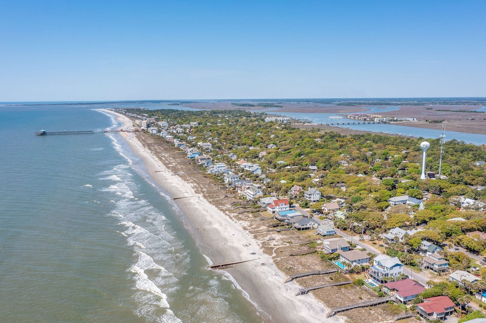 A sweeping aerial view of Folly Beach, South Carolina, reveals the island's full length of sandy shoreline, colorful beach houses, the iconic fishing pier, and the Folly Beach water tower, flanked by the Atlantic Ocean on one side and tidal marshes on the other.
