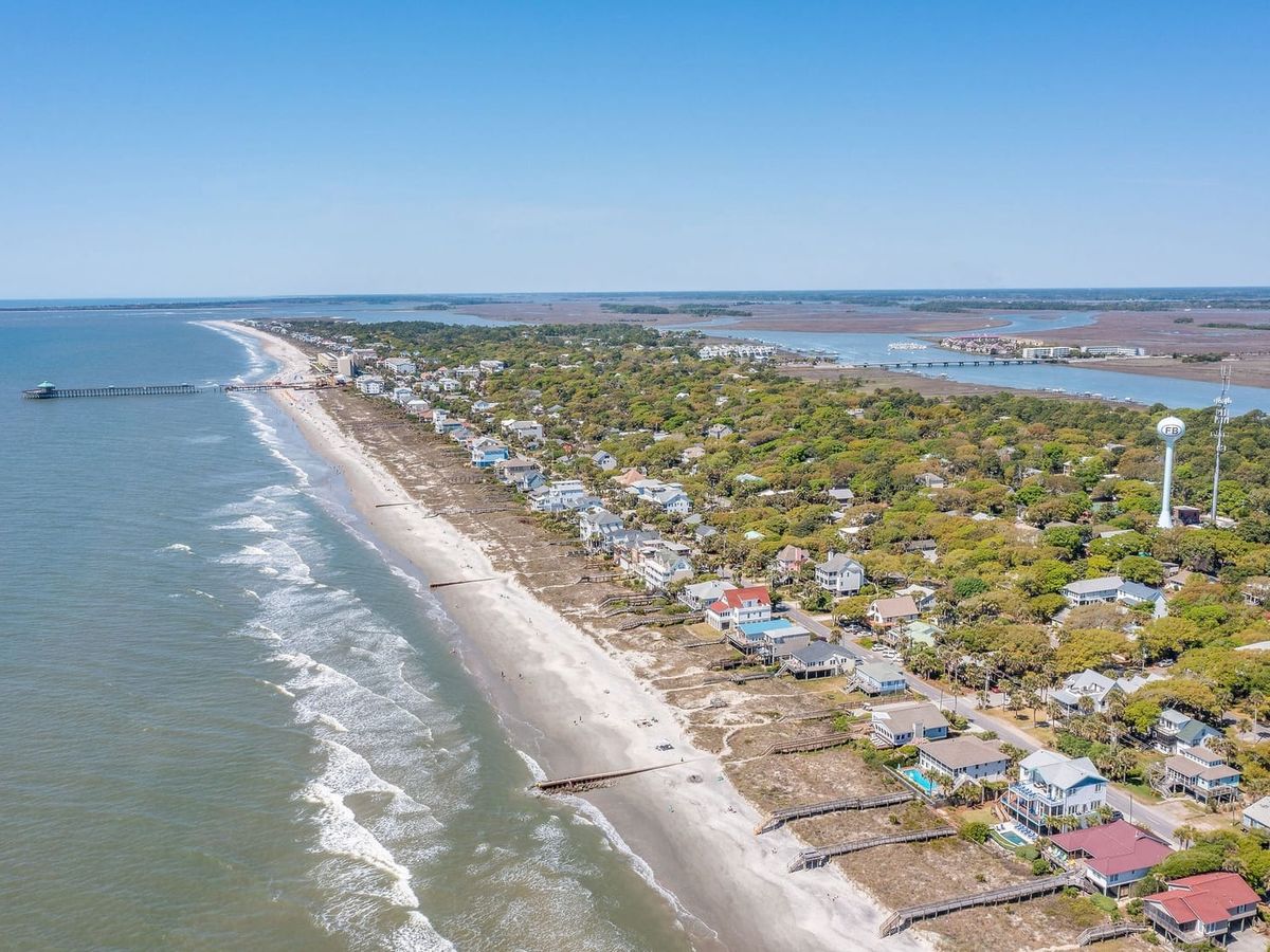 A sweeping aerial view of Folly Beach, South Carolina, reveals the island's full length of sandy shoreline, colorful beach houses, the iconic fishing pier, and the Folly Beach water tower, flanked by the Atlantic Ocean on one side and tidal marshes on the other.