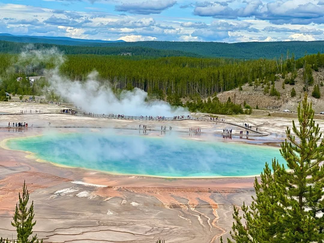 The Grand Prismatic Spring in Yellowstone shows off vibrant colors of blue, green, and orange with steam rising into the sky. Visitors walk along the boardwalk, taking in one of the park’s most iconic geothermal wonders.