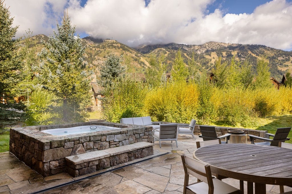 Outdoor stone hot tub set on a mountain patio with lounge seating and dining table, surrounded by golden autumn foliage and evergreen trees, with dramatic mountain peaks rising in the background.