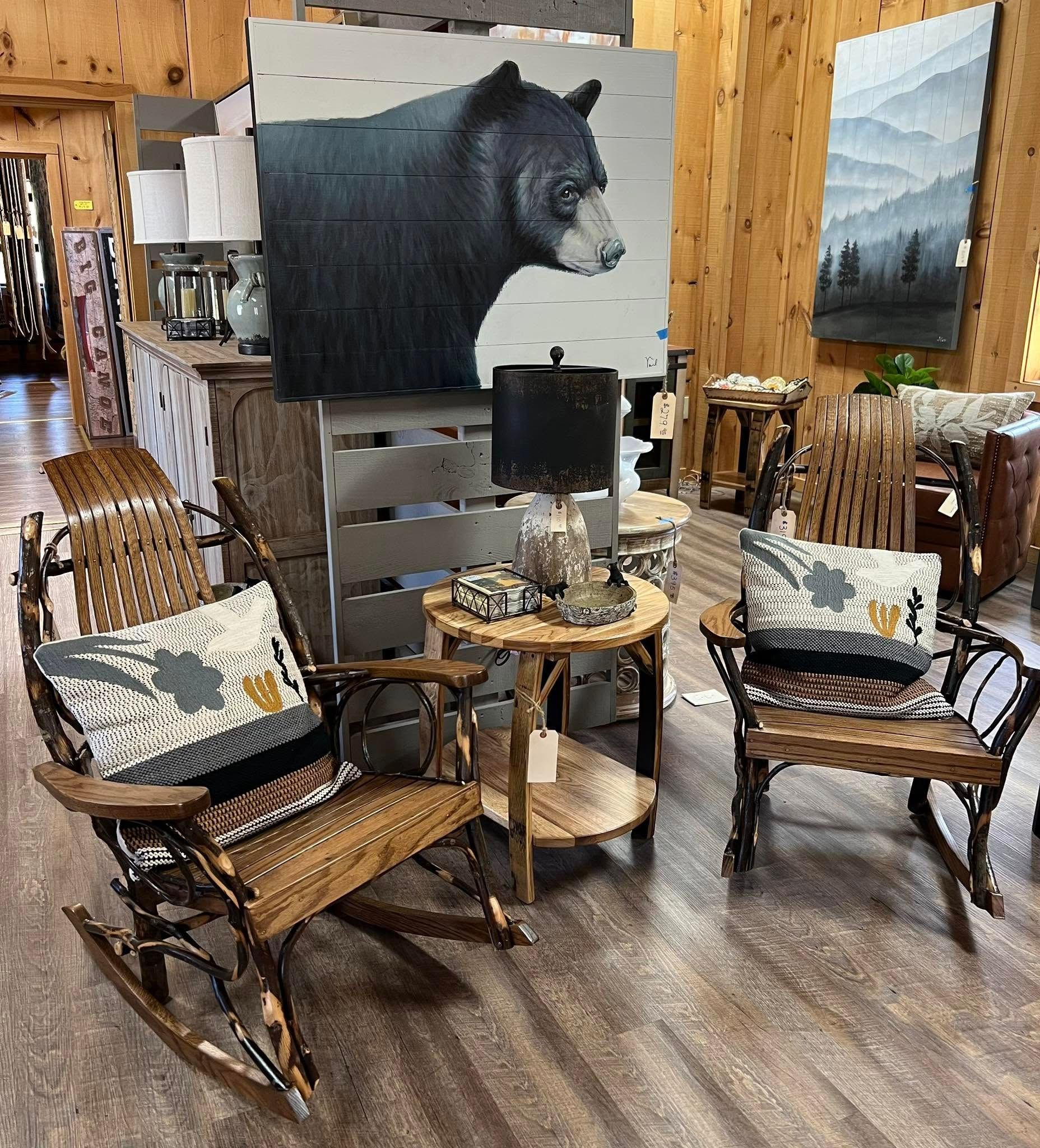 A rustic furniture display at Lazy Bear Furniture in Georgia featuring two handcrafted wooden rocking chairs with patterned pillows, a small round side table, and a cozy lamp. Behind the setup, a striking bear painting and wood-paneled walls add to the warm mountain cabin atmosphere.