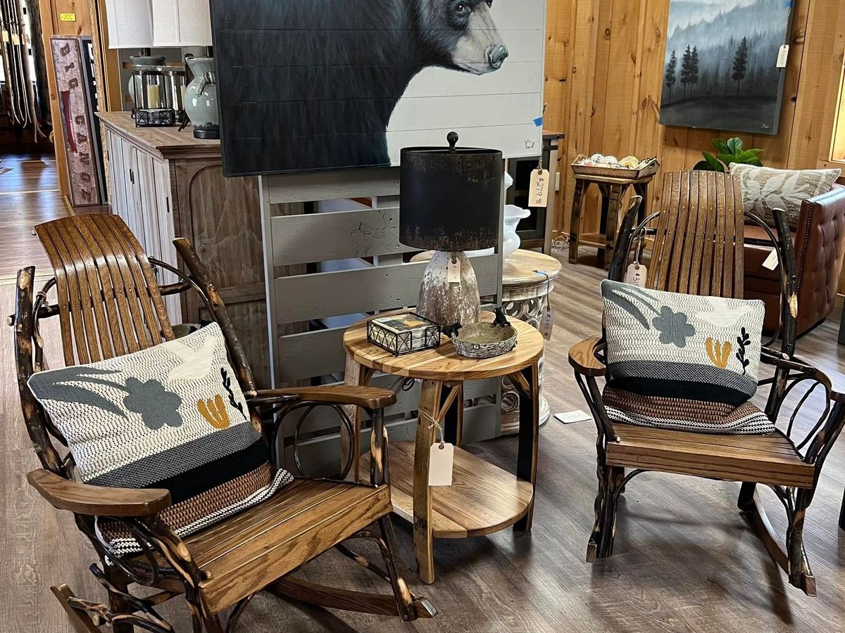 A rustic furniture display at Lazy Bear Furniture in Georgia featuring two handcrafted wooden rocking chairs with patterned pillows, a small round side table, and a cozy lamp. Behind the setup, a striking bear painting and wood-paneled walls add to the warm mountain cabin atmosphere.