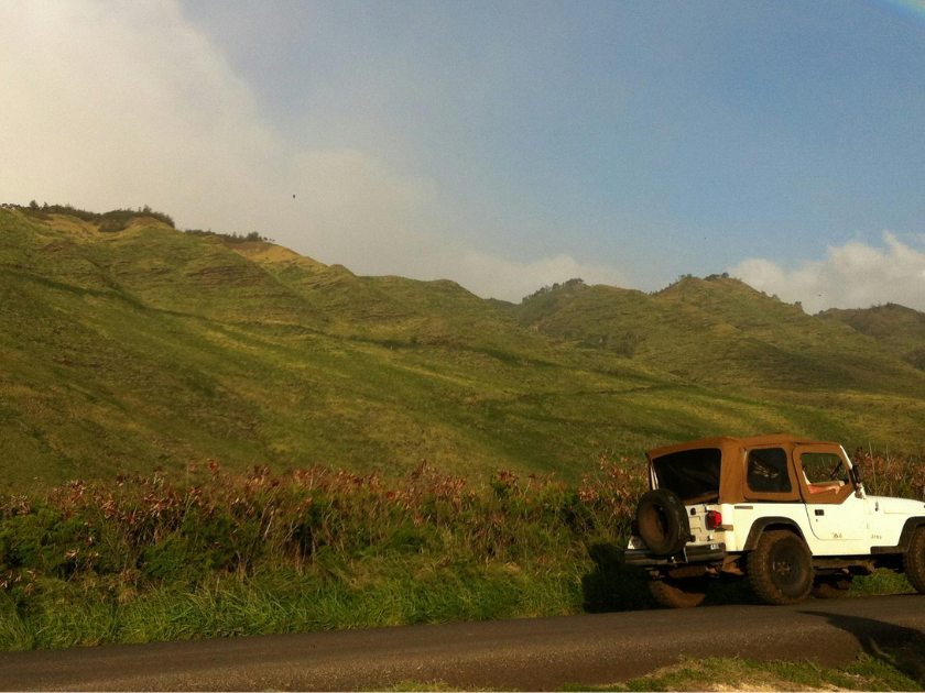 A white Jeep parked on the side of a road in front of green hills and a rainbow. Exploring Hawaii by car lets you reach remote spots and chase sunsets on your own schedule.