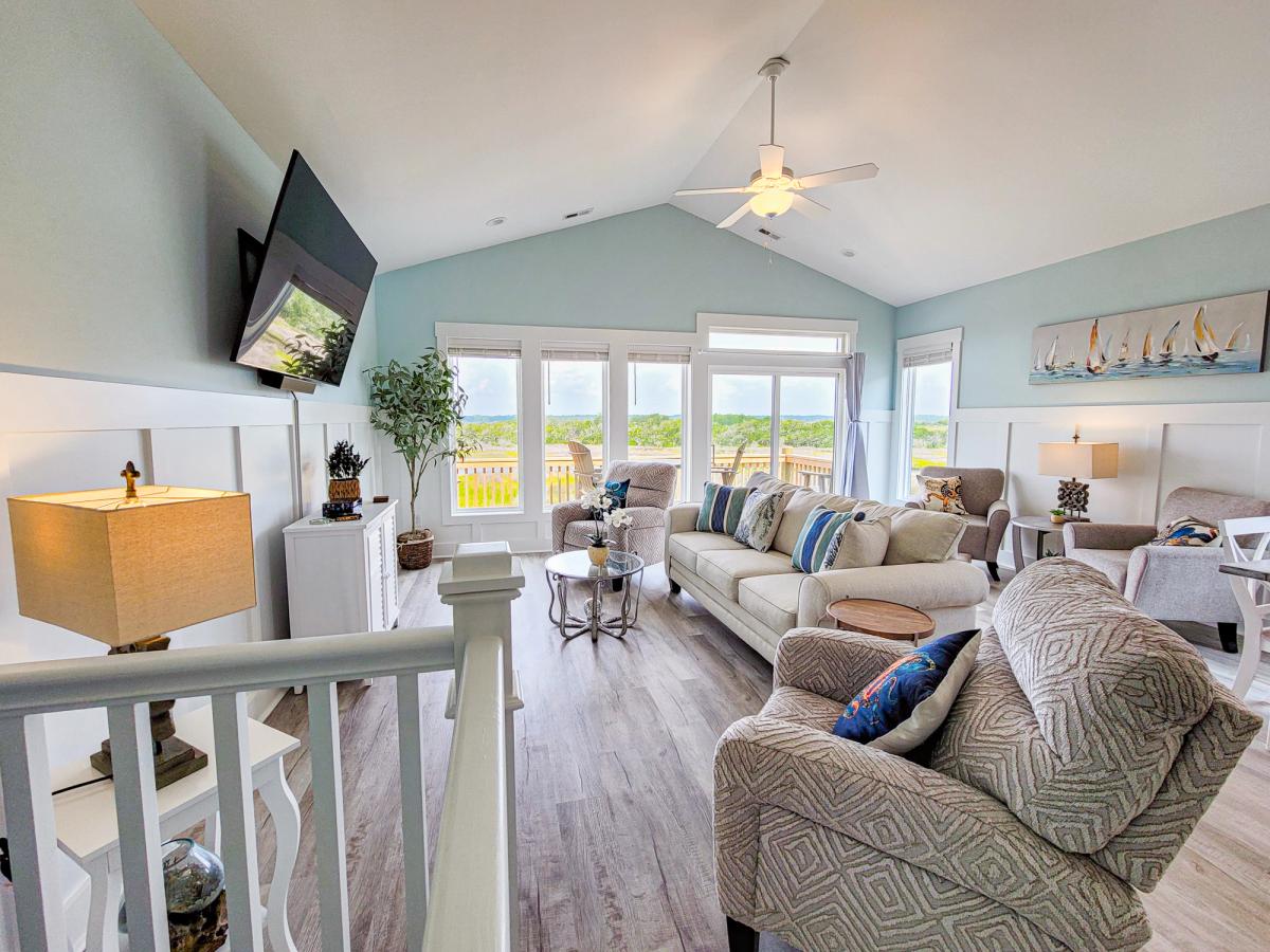 A bright and airy coastal living room with vaulted ceilings, light blue walls, and large windows letting in natural light, furnished with neutral-toned sofas, patterned armchairs, beach-themed decor, and a wall-mounted TV.