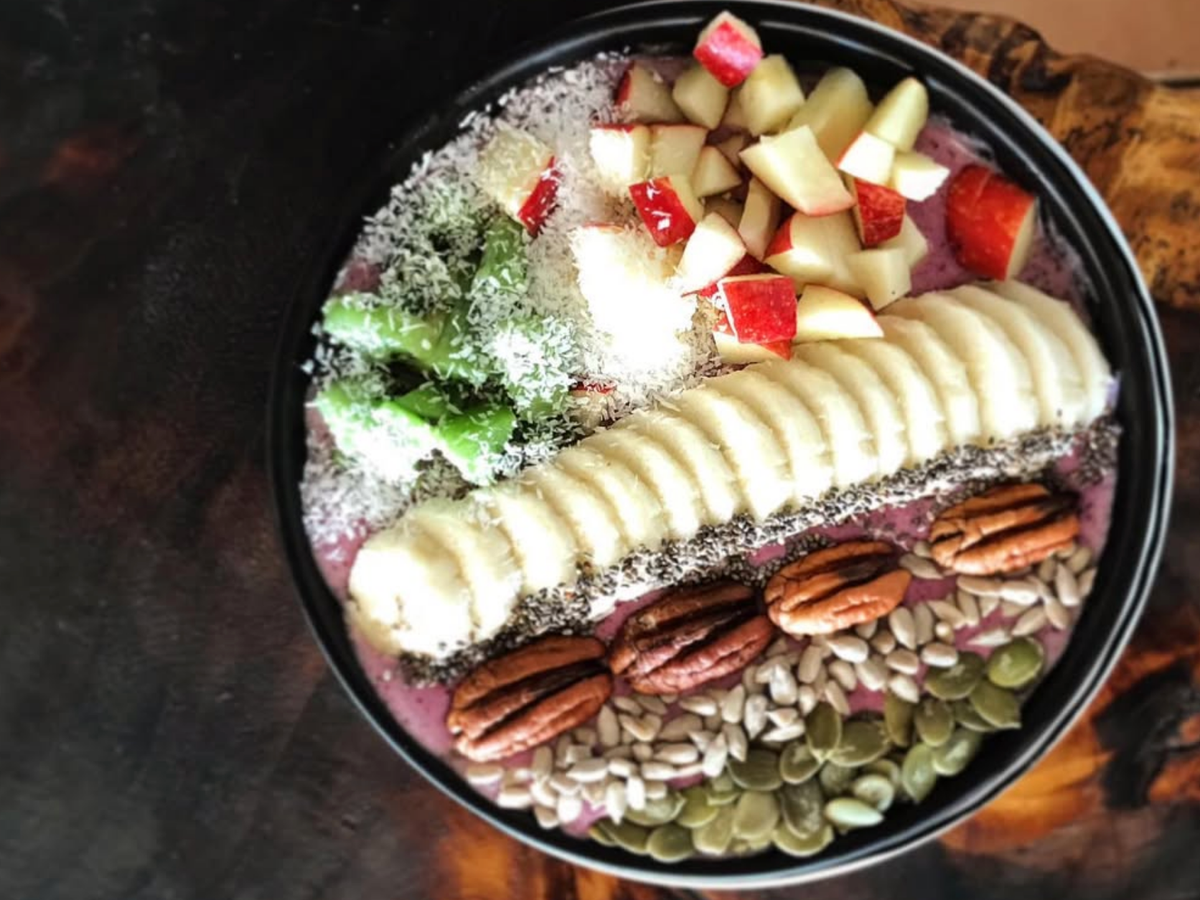 A colorful smoothie bowl topped with sliced banana, diced apples, shredded coconut, chia seeds, pecans, sunflower seeds, pumpkin seeds, and fresh greens, served in a black bowl on a dark wooden surface.